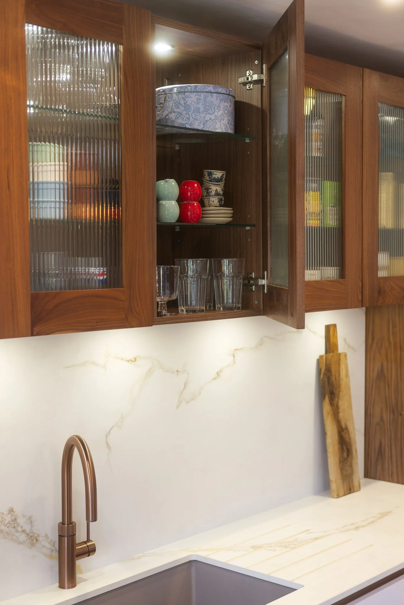 Open wooden kitchen cabinet with glass doors showing bowls, cups, and glasses inside, above a white marble countertop with a gold faucet and a leaning wooden cutting board.