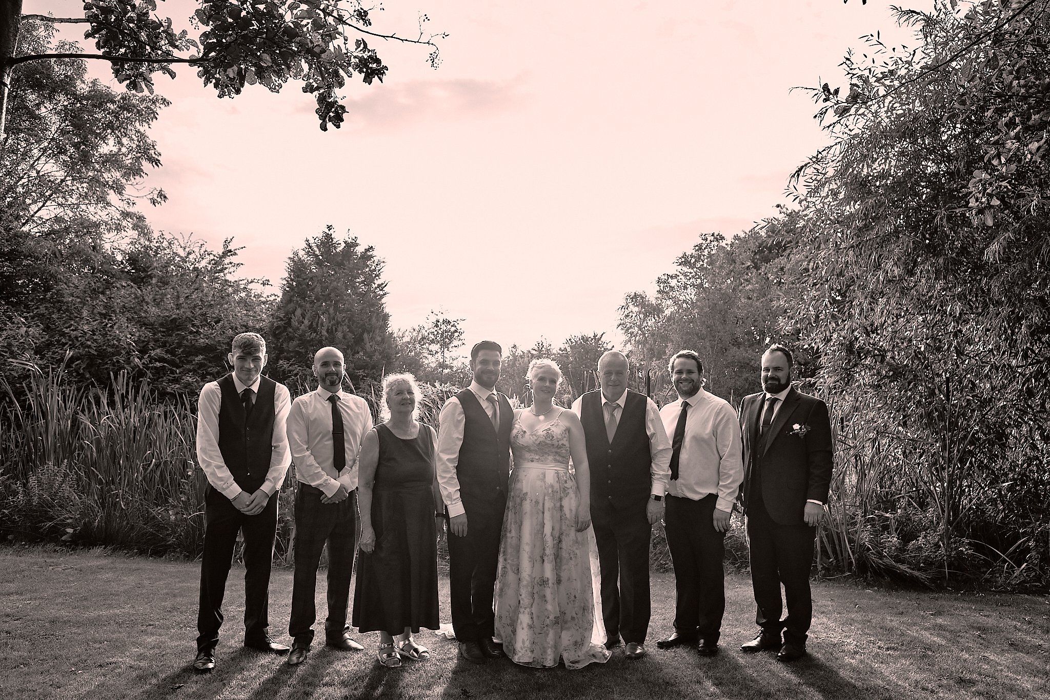 A group of nine people, including a bride in a long floral dress and a groom in a suit, standing outdoors in front of trees and bushes. It appears to be a wedding photo taken during sunset.
