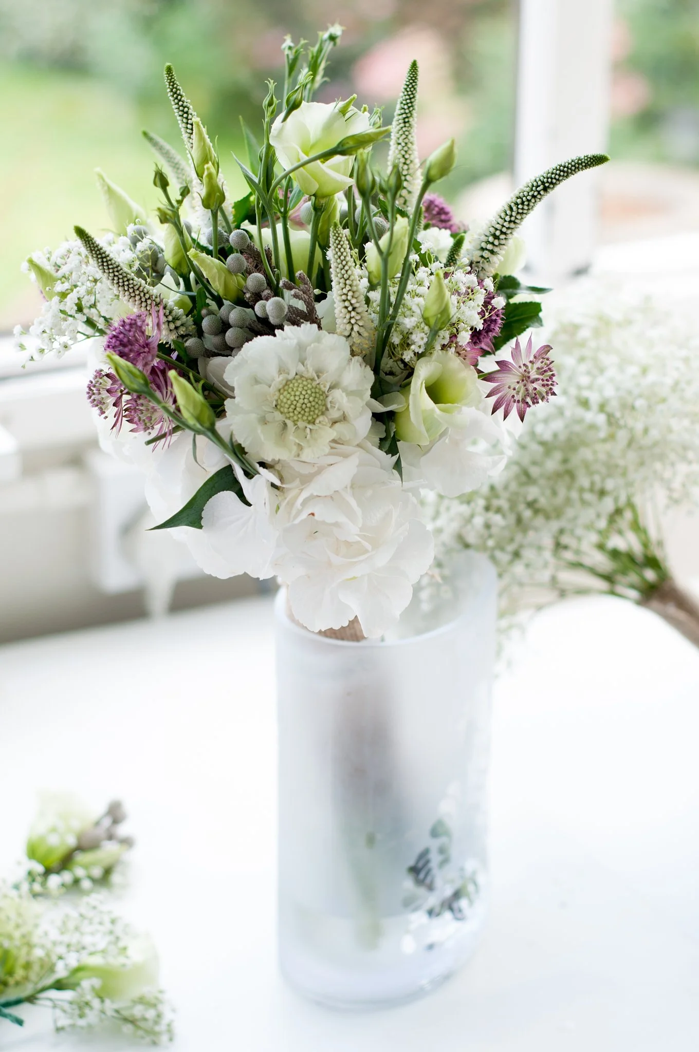 A tall white vase with a floral arrangement of white, purple, and green flowers, with a blurred background of greenery and a window.