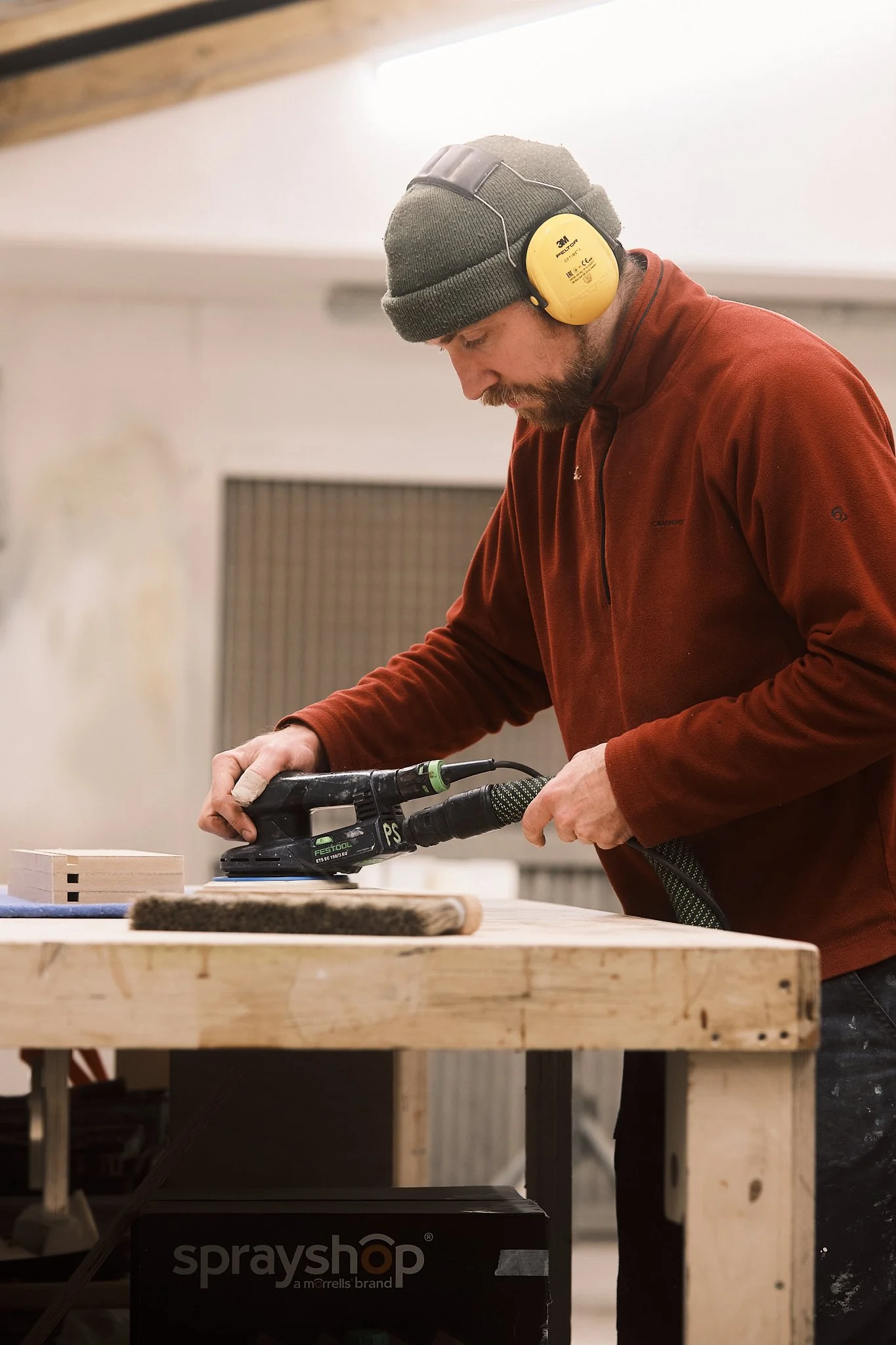 A man wearing a gray beanie, yellow hearing protection, and a red fleece jacket is sanding a piece of wood with a handheld sander in a woodworking shop.