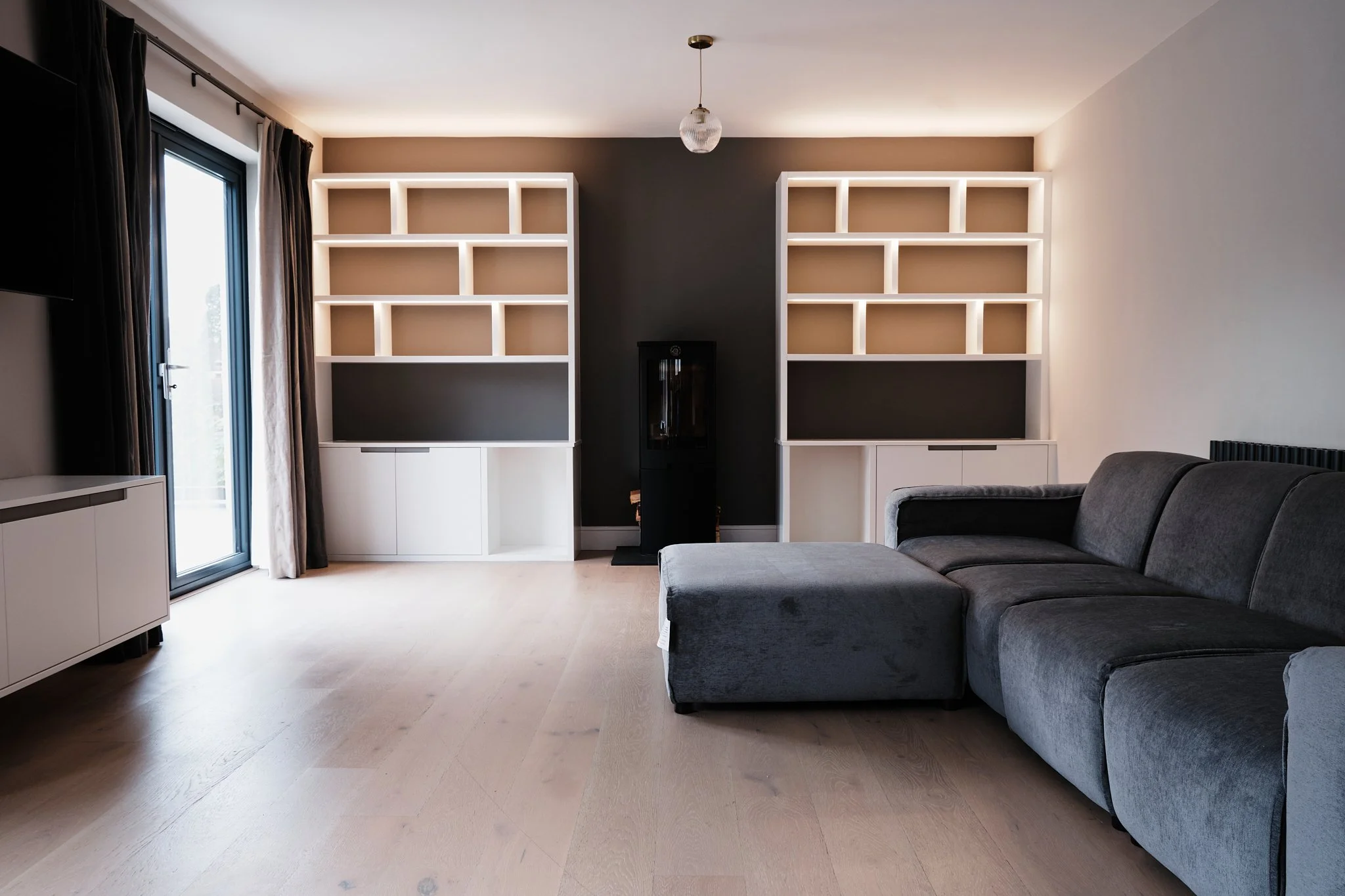 Modern living room with a dark gray sectional sofa, light wood flooring, two large, white built-in bookshelves, a black water cooler in the middle, and a glass door with curtains on the left.