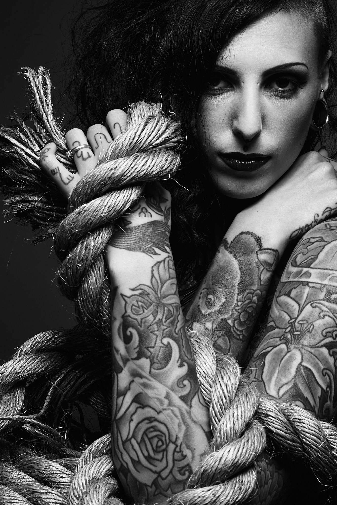 Black and white portrait of a woman with tattoos, dark makeup, and dark curly hair, holding thick rope around her shoulders.