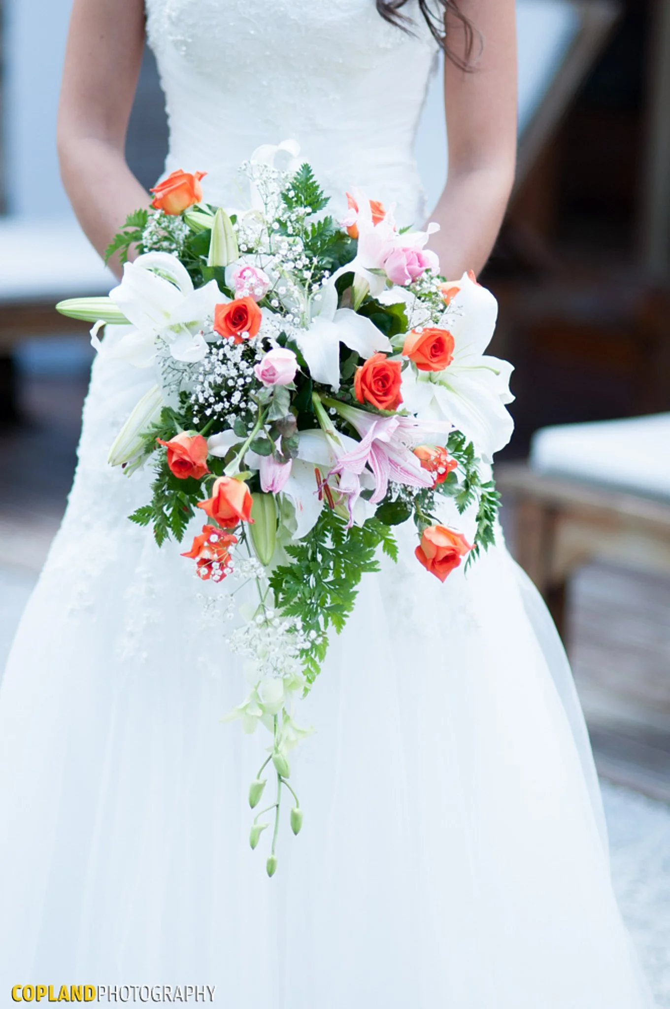 Wedding dress with a cascading bouquet of white lilies, orange roses, pink roses, baby's breath, and various greenery.