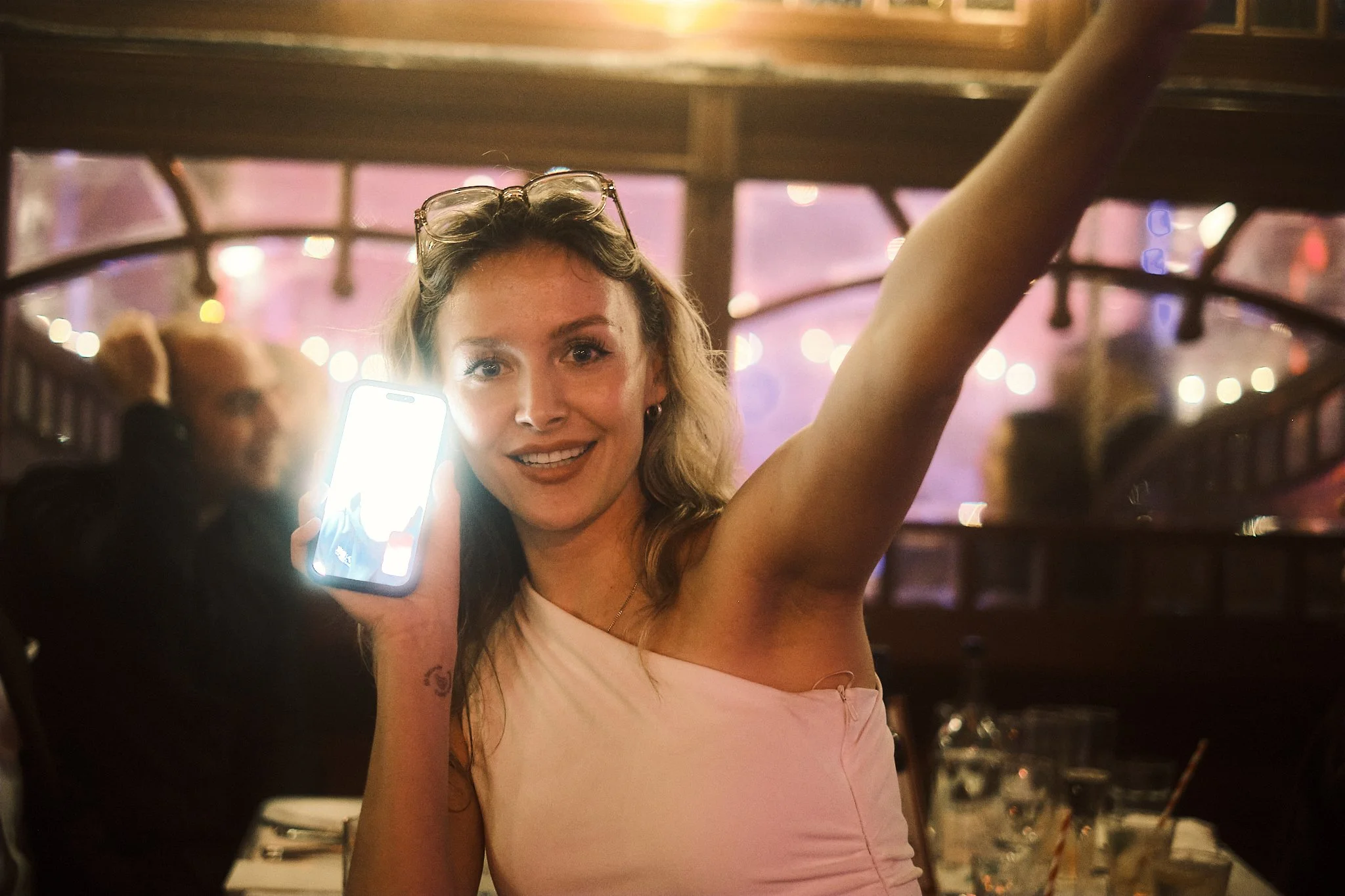 A woman at a restaurant holding a smartphone, smiling, with glasses on her head, in a lively indoor setting with warm lighting and other people in the background.