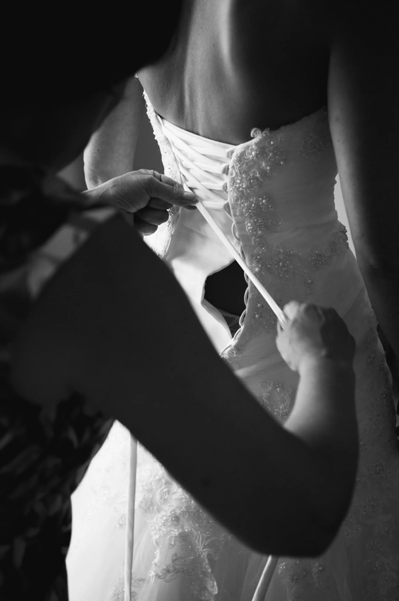 Close-up black-and-white photo of a person in a wedding dress with lace details, as someone adjusts the back with a ribbon in a dimly lit setting.