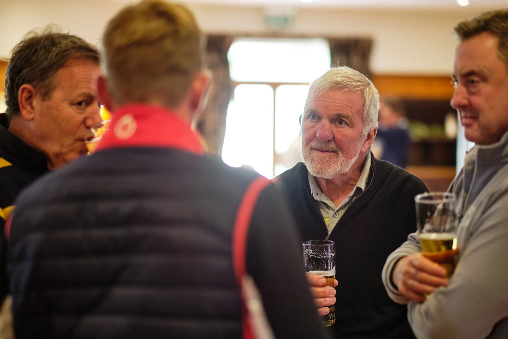 Four men engaged in conversation at a social gathering, holding glasses of beer, indoors with natural light coming through a window in the background.