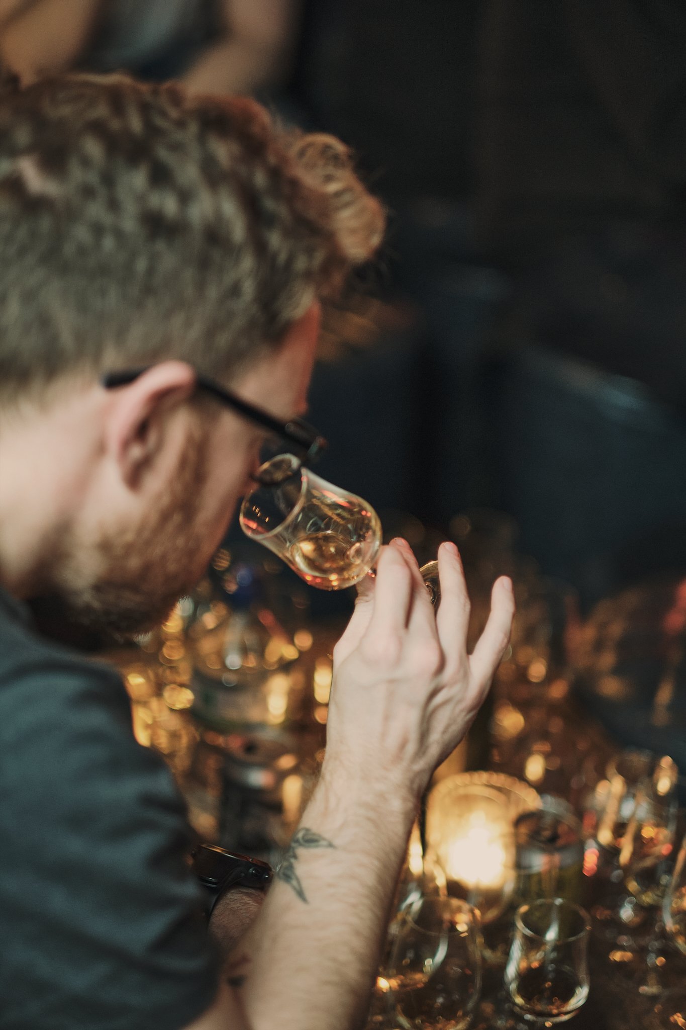 A man with glasses and a tattoo on his forearm enjoying a glass of whiskey at a bar with dim lighting and multiple glasses in the background.