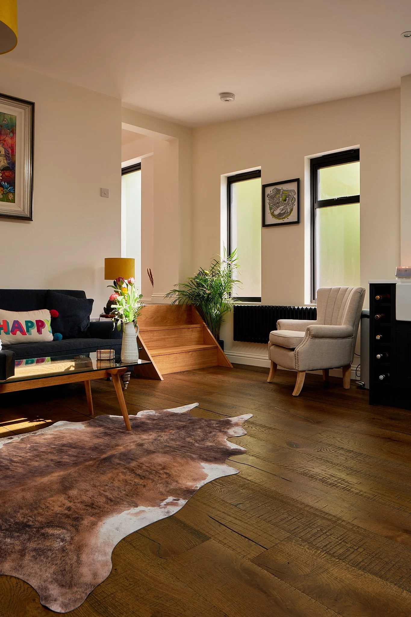 Living room with wooden floors, a cowhide rug, a black sofa with colorful pillows, a glass coffee table, a beige armchair, plants near windows, and framed artwork on walls.