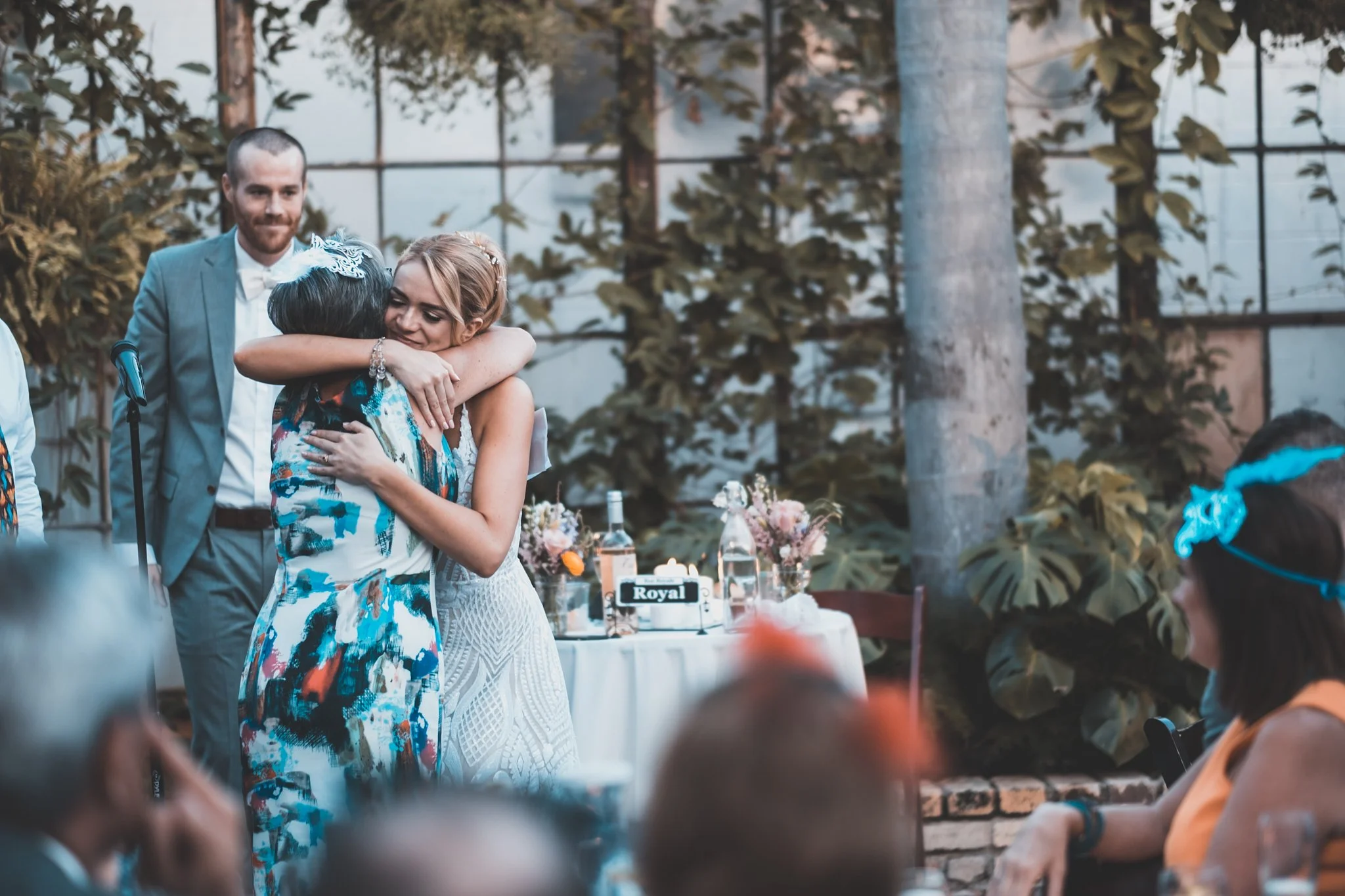 A bride and an older woman hugging at a wedding reception, with a groom standing nearby. The scene is outdoors with trees and tables with floral arrangements and bottled drinks visible.