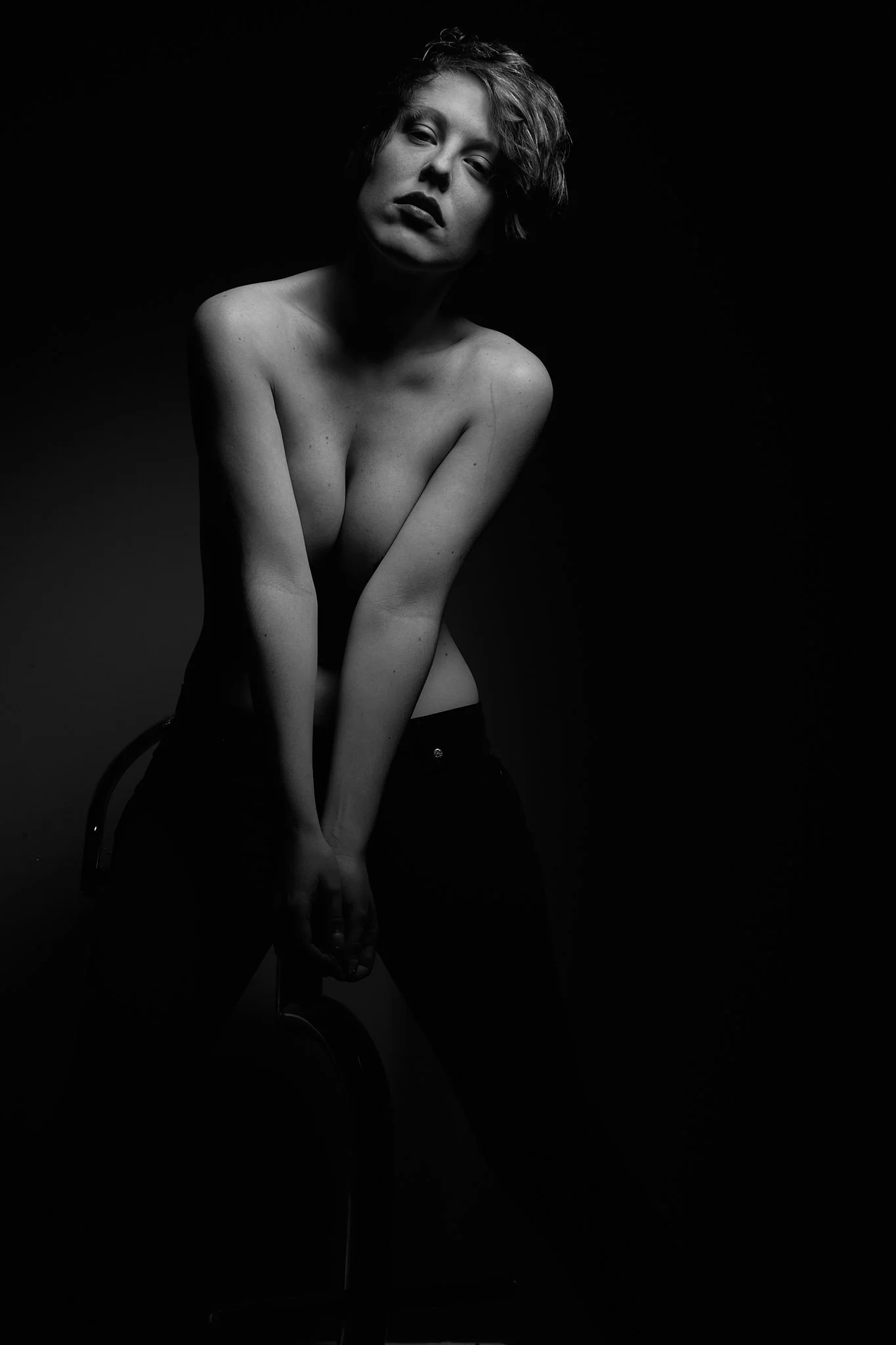 Black and white photograph of a woman with short hair, topless, sitting on a chair in low lighting, with her hands clasped in front of her.