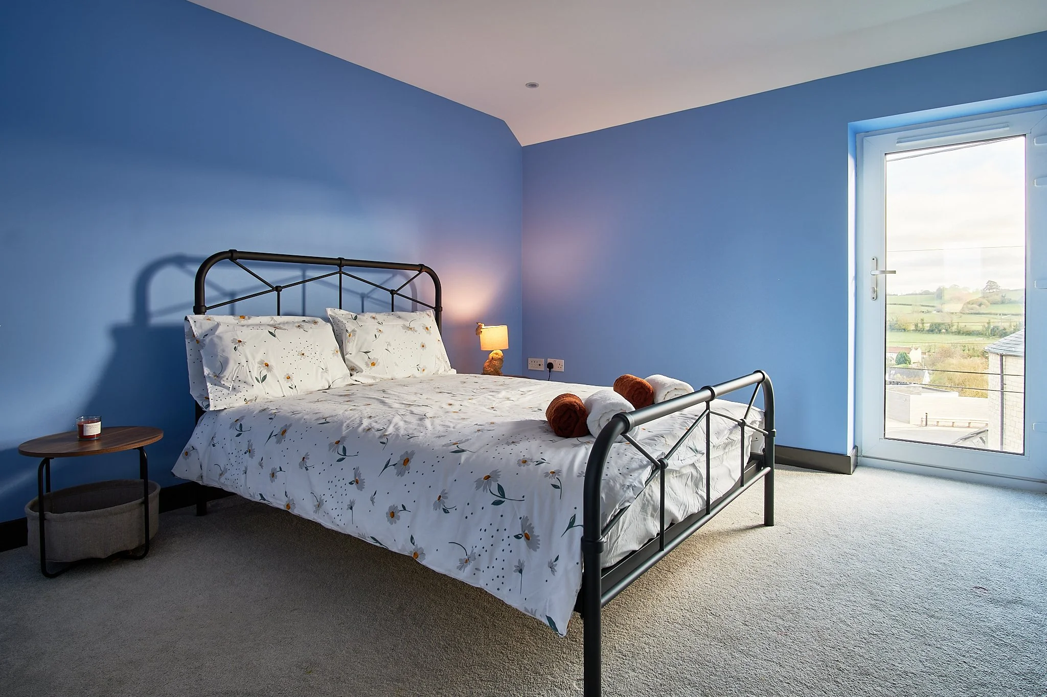 A bedroom with blue walls, a black metal bed frame with white floral bedding, a small round wooden bedside table with a candle, a lit lamp, and rolled towels, a large window showing a view of fields and houses.