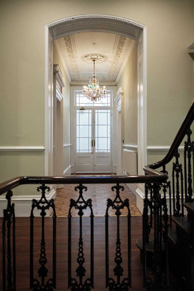 View of an elegant entryway with a chandelier, a decorative ceiling, and a large front door with glass panels, taken from the staircase landing with ornate black iron railings.