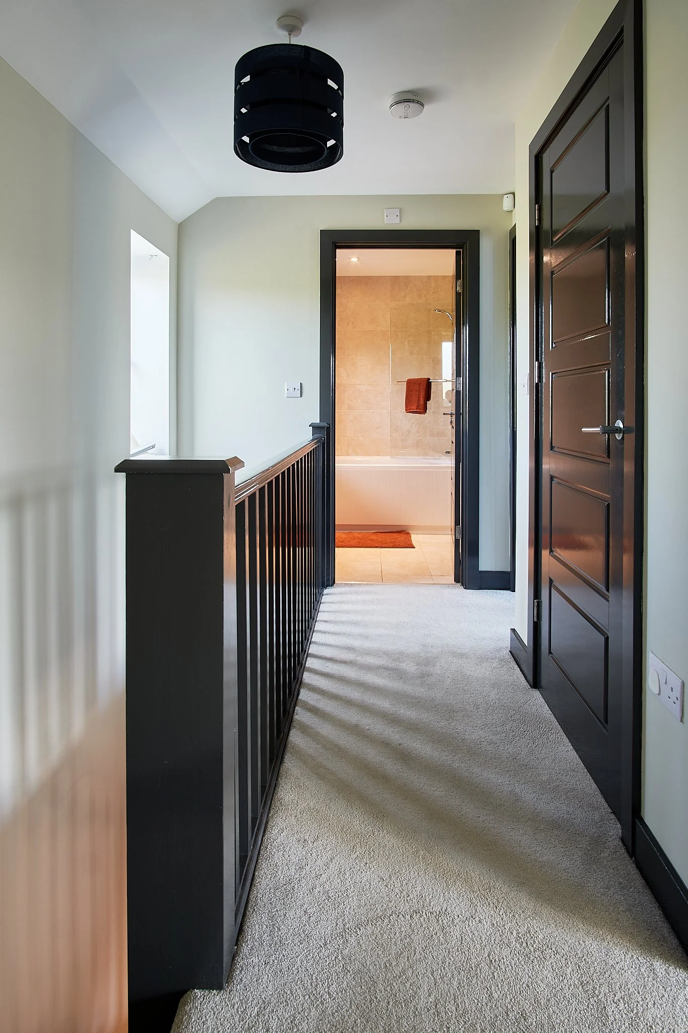 A hallway with beige carpet, black railing, black door, light green walls, leading to a bathroom with beige tiled walls, a bathtub, and an orange towel hanging on a rail.