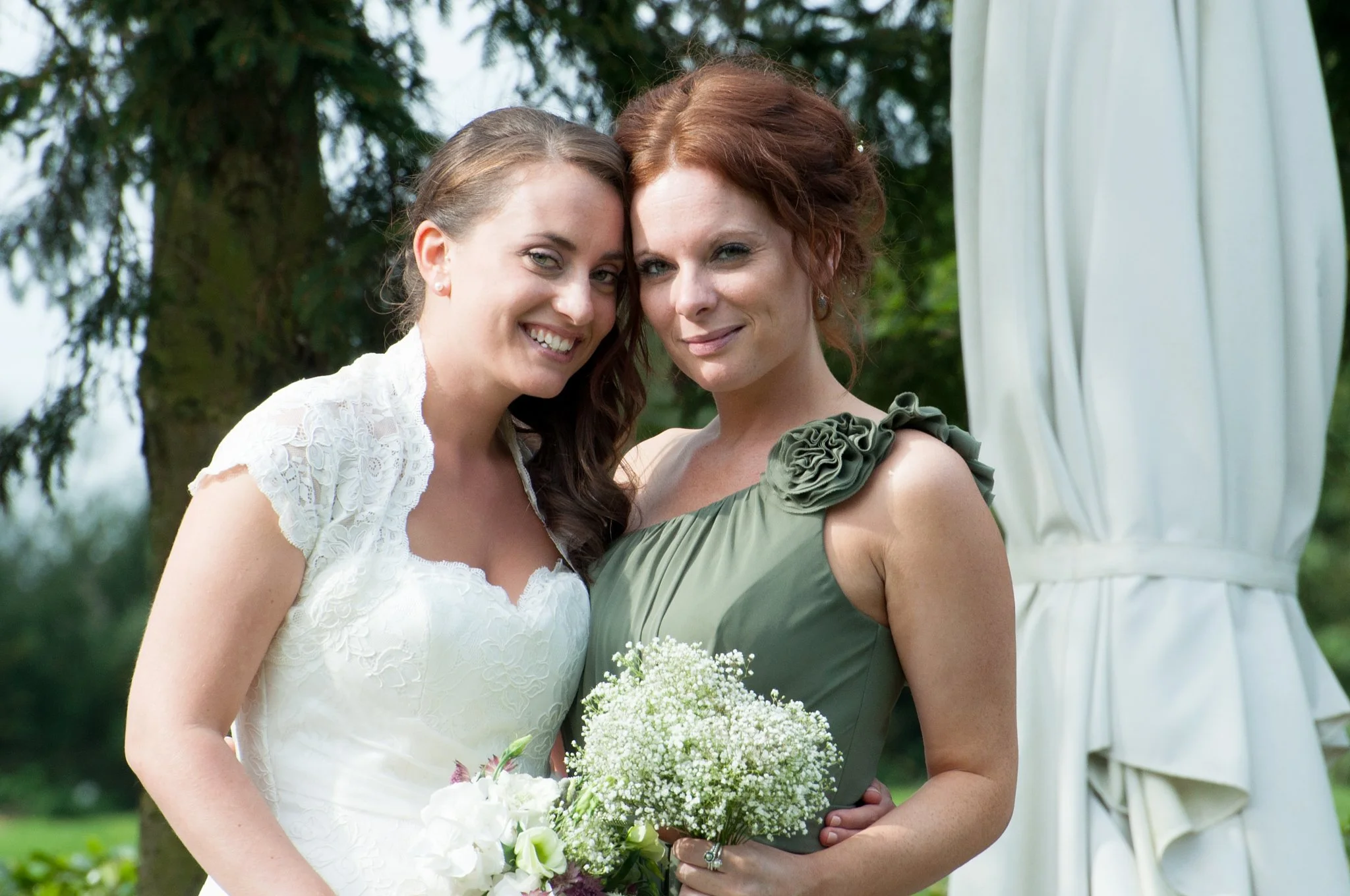 Two women at a wedding, posing close together outdoors, with one in a white wedding dress holding a bouquet, and the other in a green dress holding a smaller bouquet, both smiling and leaning their heads together.