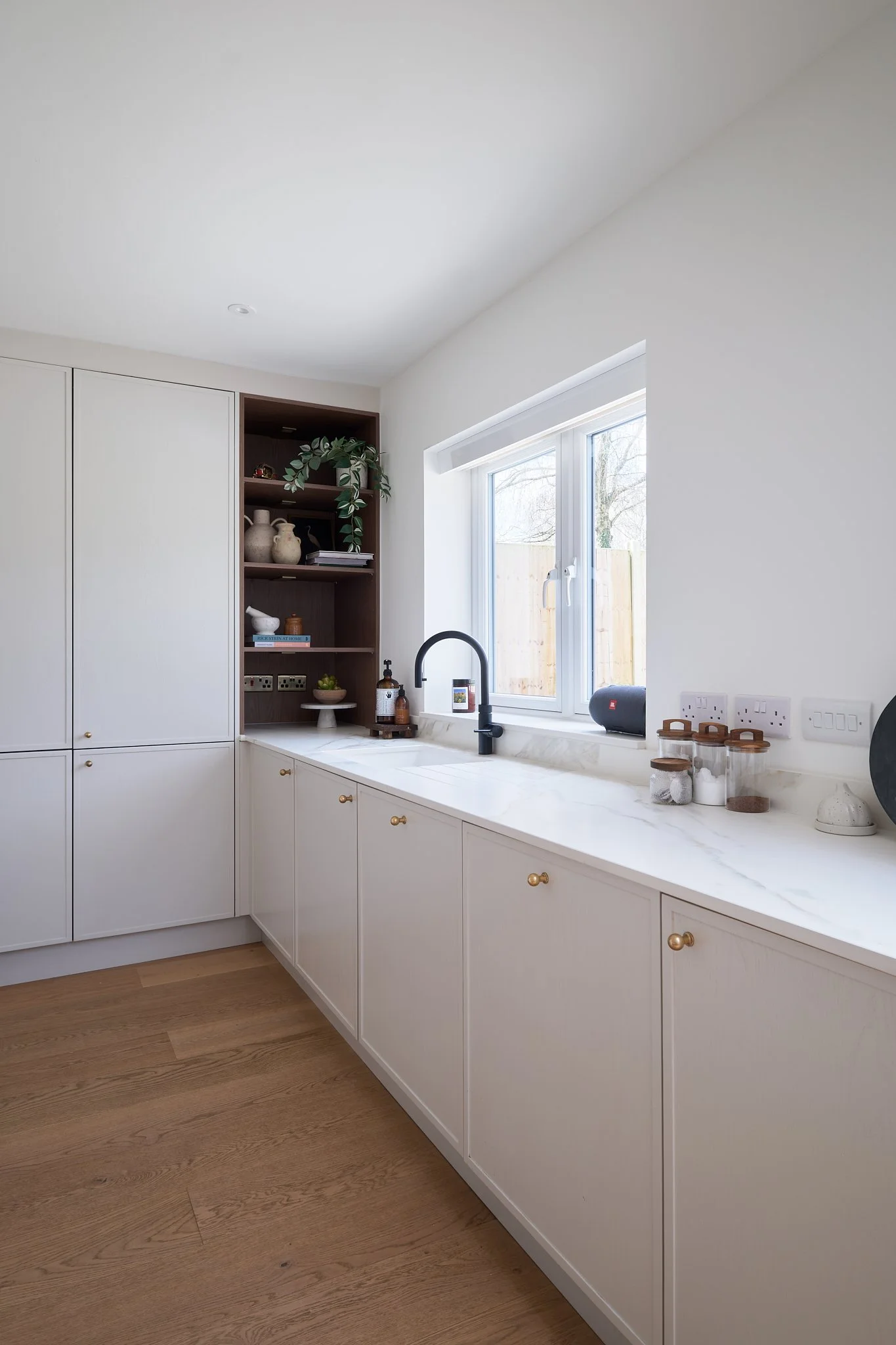 A clean, modern kitchen with white cabinets, a marble countertop, and a window with natural light. There are small decor items, jars, and a black sink with a black faucet.