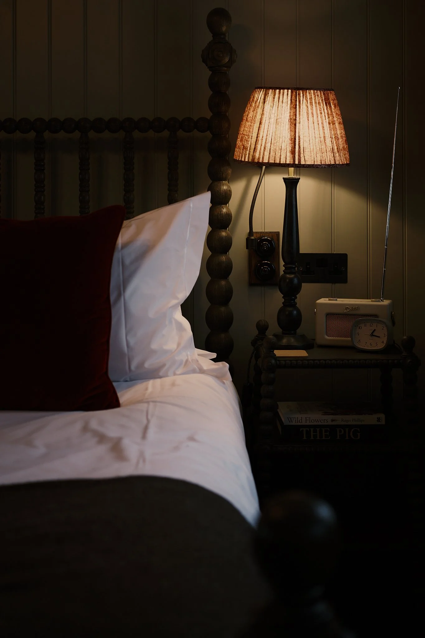 A cozy bedroom corner with a bed, white and red pillows, a bedside table, a lamp, a vintage radio, a clock, and books, illuminated by warm light. Hotel interior photography at THE PIG-in the Cotswolds