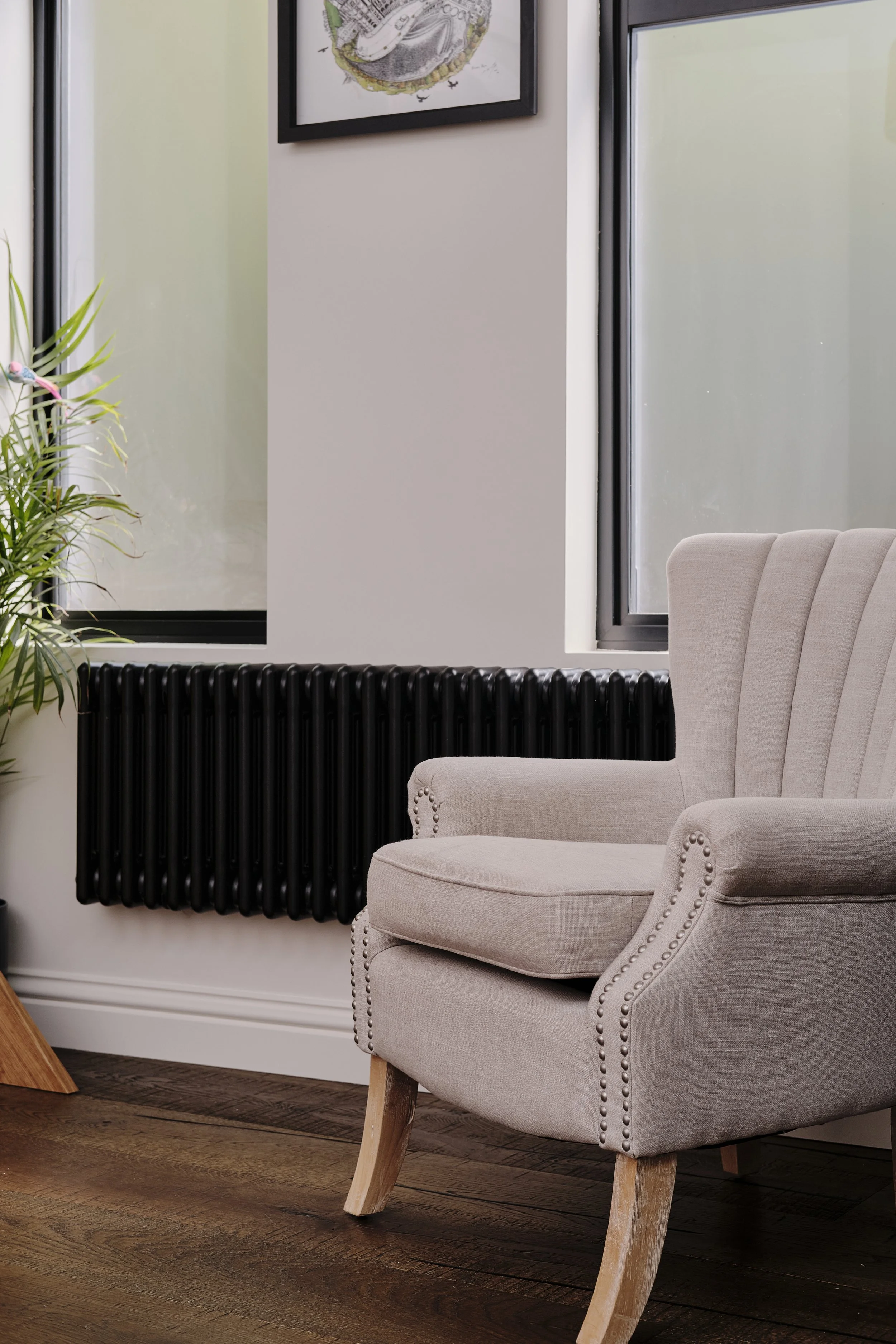 A beige upholstered armchair with wooden legs, positioned next to a black radiator and near tall windows, in a modern interior space.