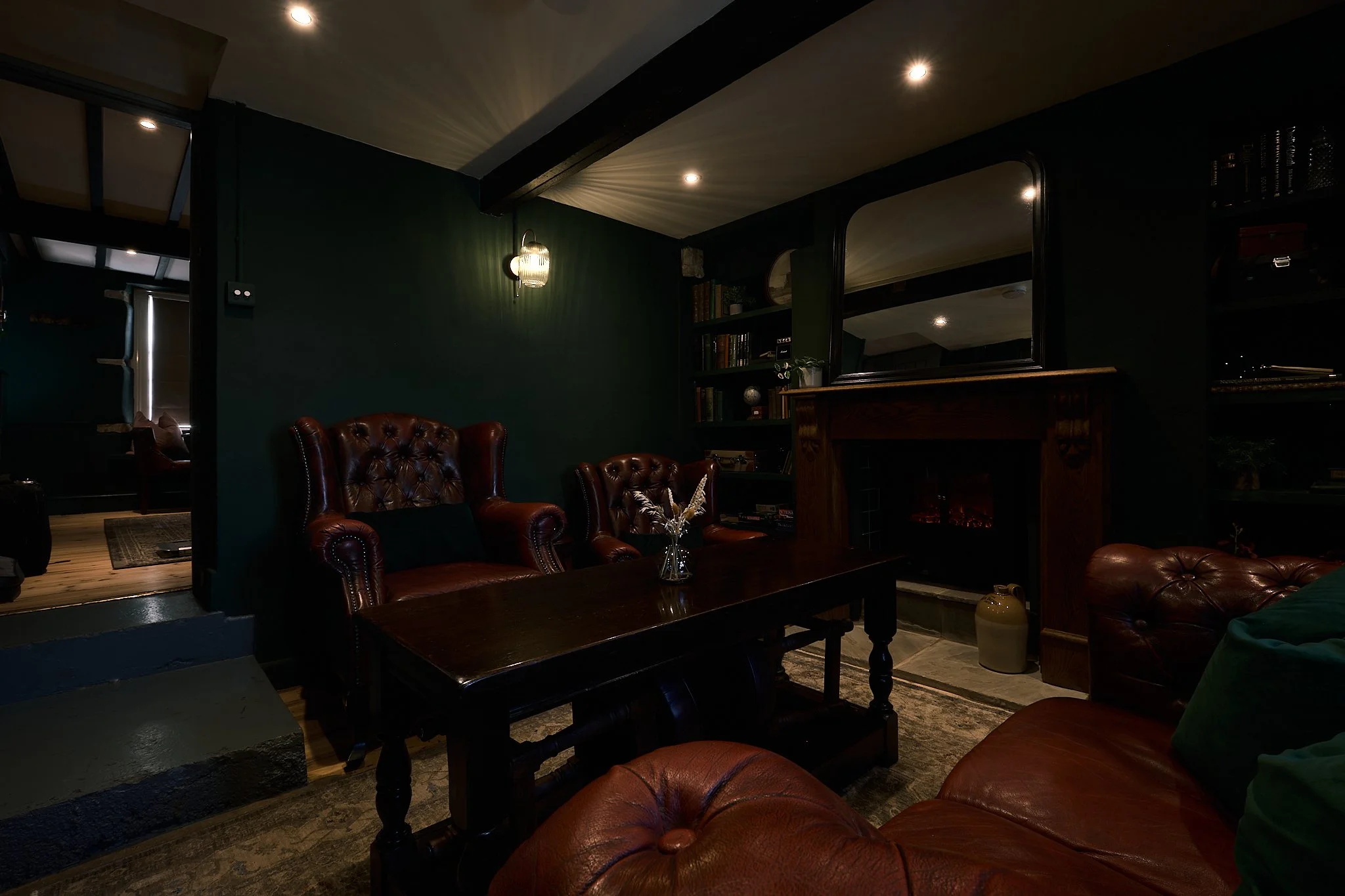 A cozy living room with dark green walls, leather armchairs, a wooden coffee table, and a fireplace, illuminated by wall sconces and recessed ceiling lights.