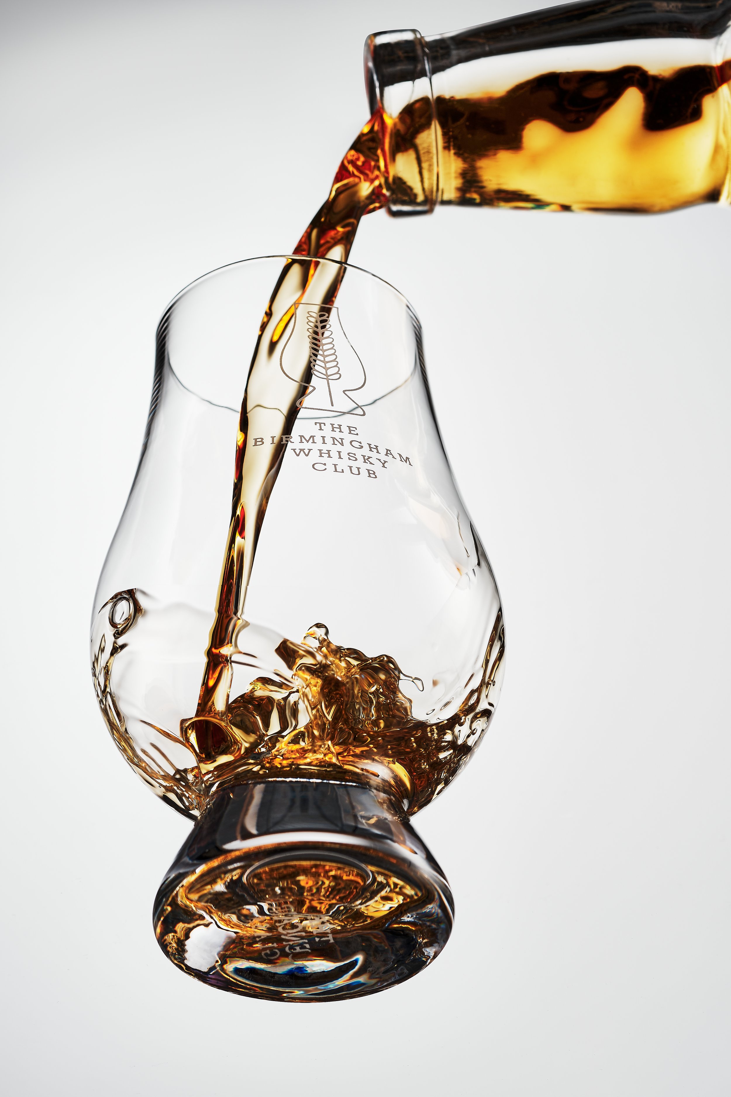 Whiskey being poured from a bottle into a branded glass with the text 'The Birmingham Whisky Club' on it, against a white background.