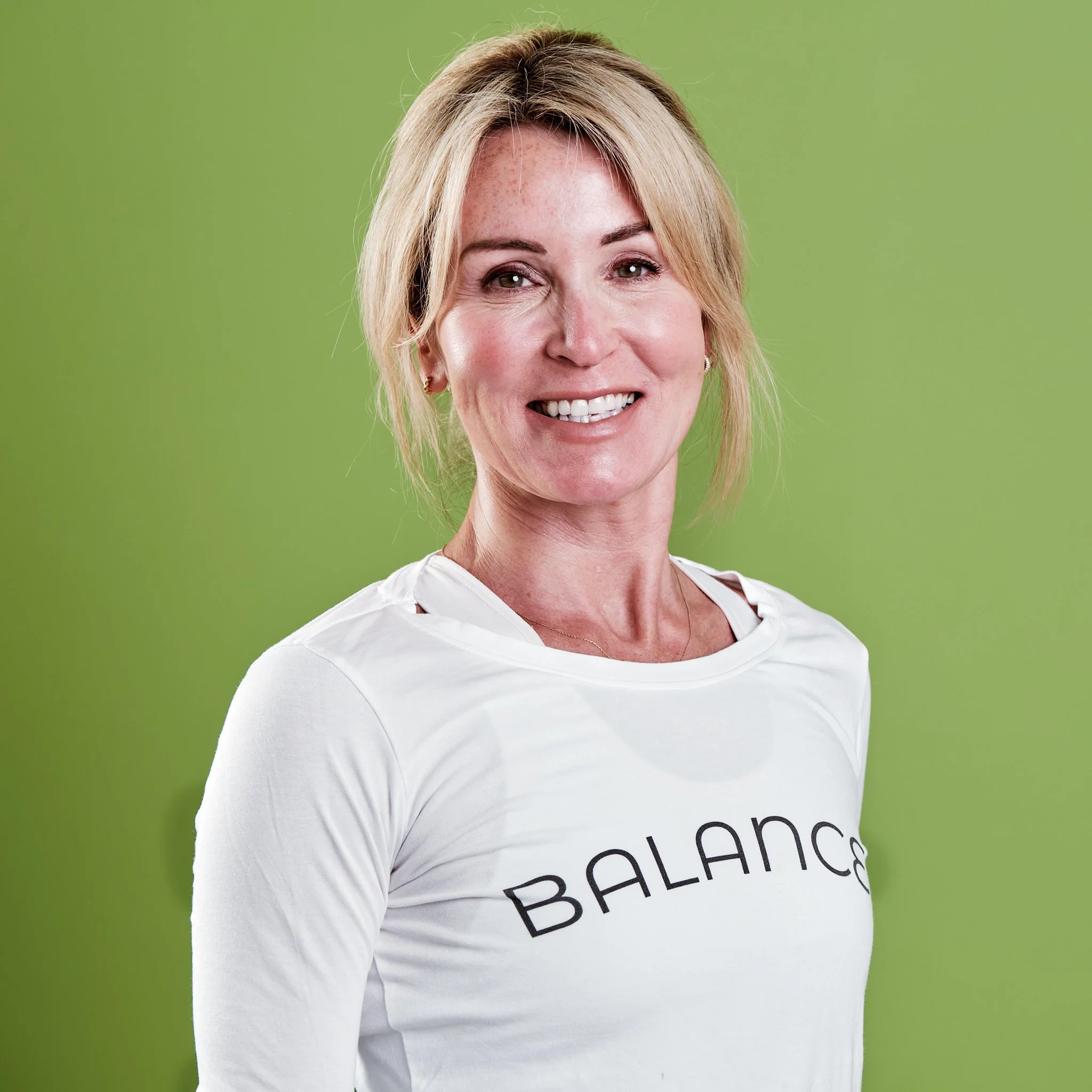 A smiling woman with blonde hair wearing a white shirt with the word "BALANCE" on it, standing against a green background.
