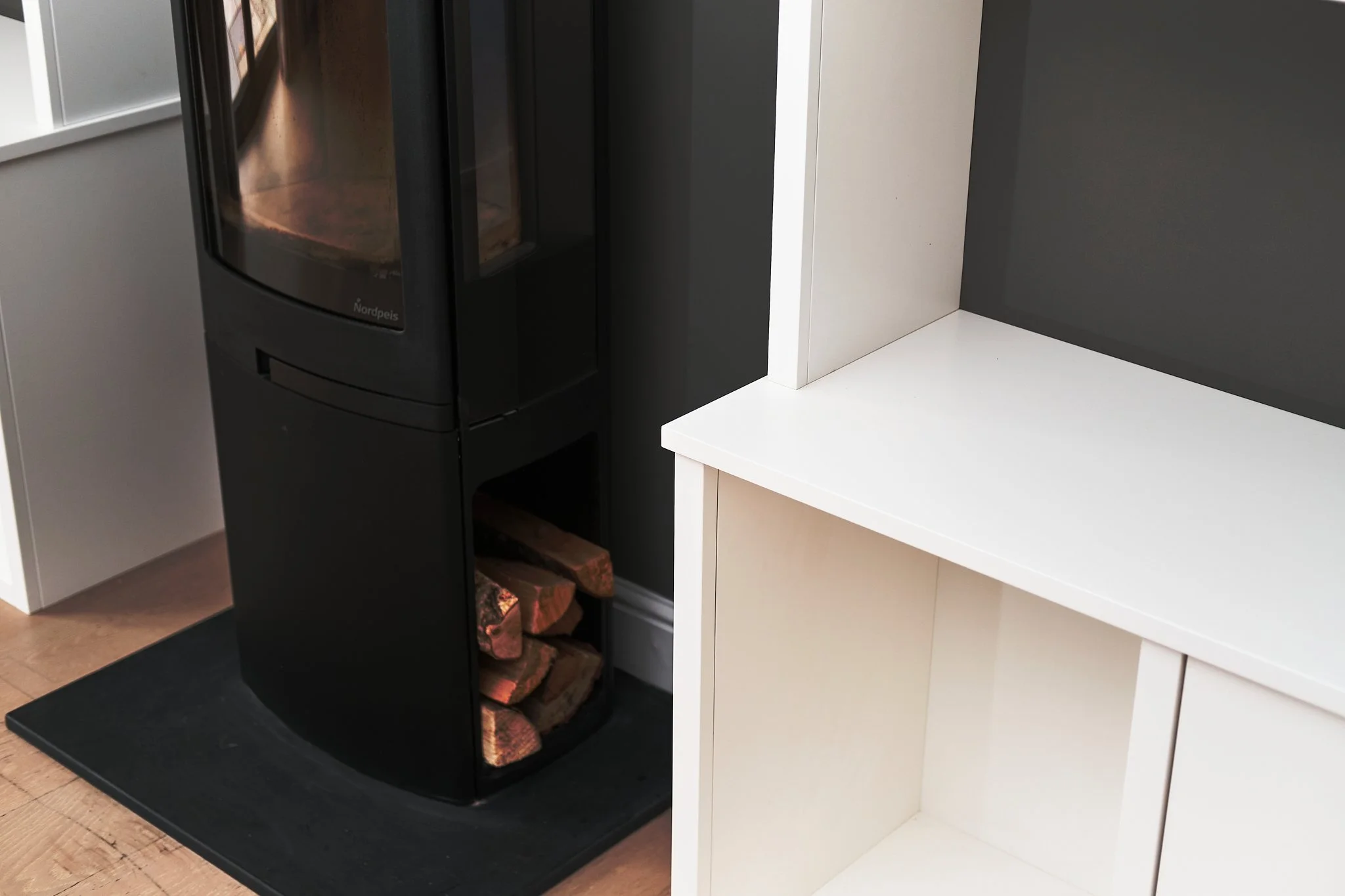 A black pellet stove with a glass door, placed on a black mat next to a white cabinet with an open shelf.