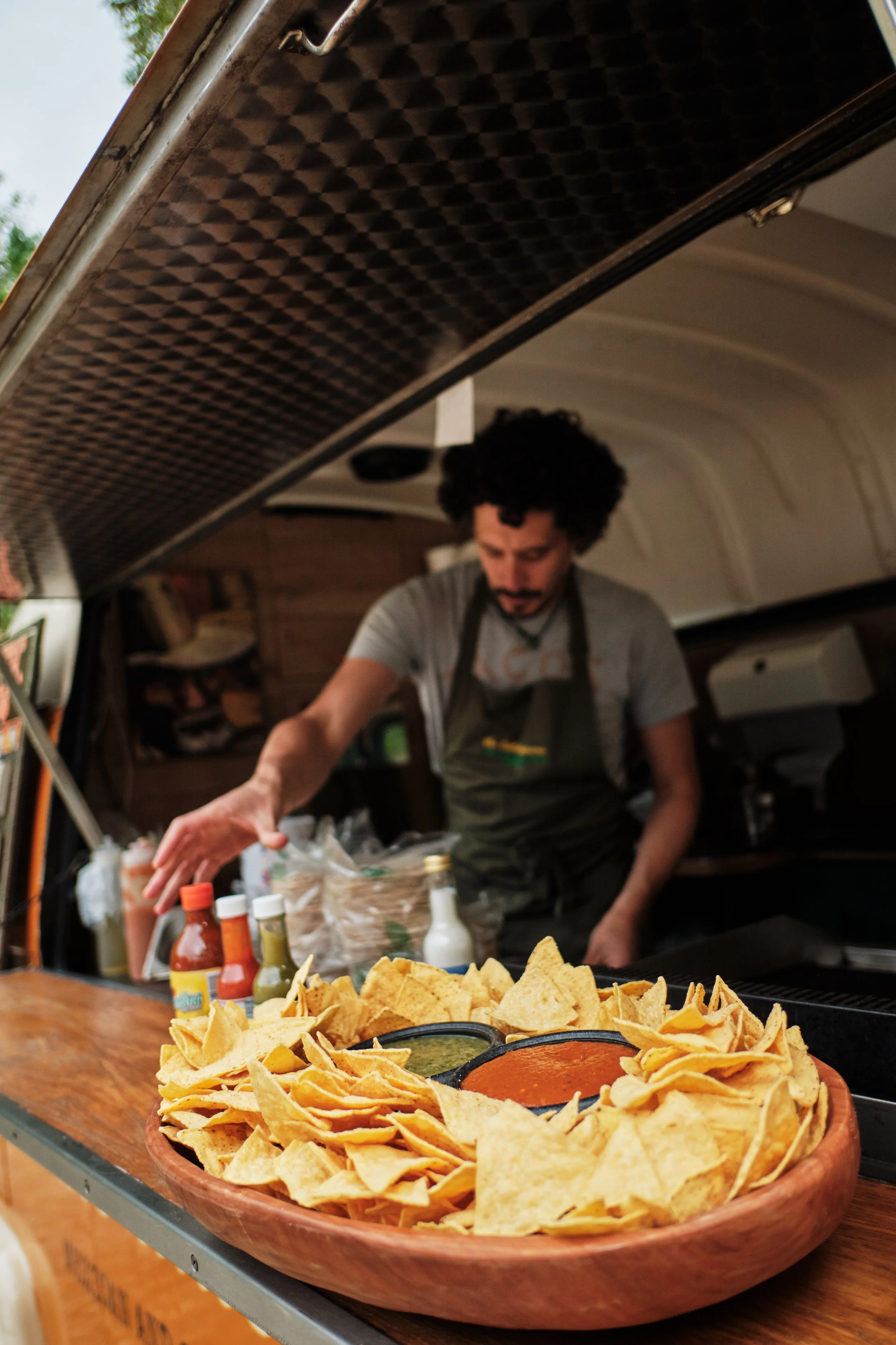 A man preparing food behind a counter with a large wooden bowl of tortilla chips and salsa in the foreground.