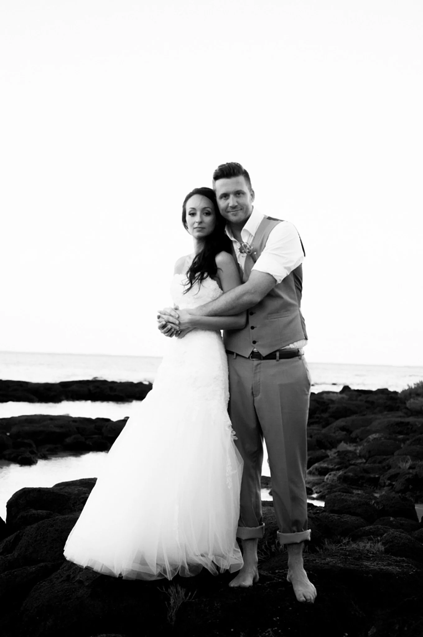 A black and white photo of a bride and groom standing on rocks near the ocean. The bride is wearing a strapless wedding dress and has long, dark hair. The groom is dressed in a vest and shirt with rolled-up pants, and is barefoot. They are embracing 