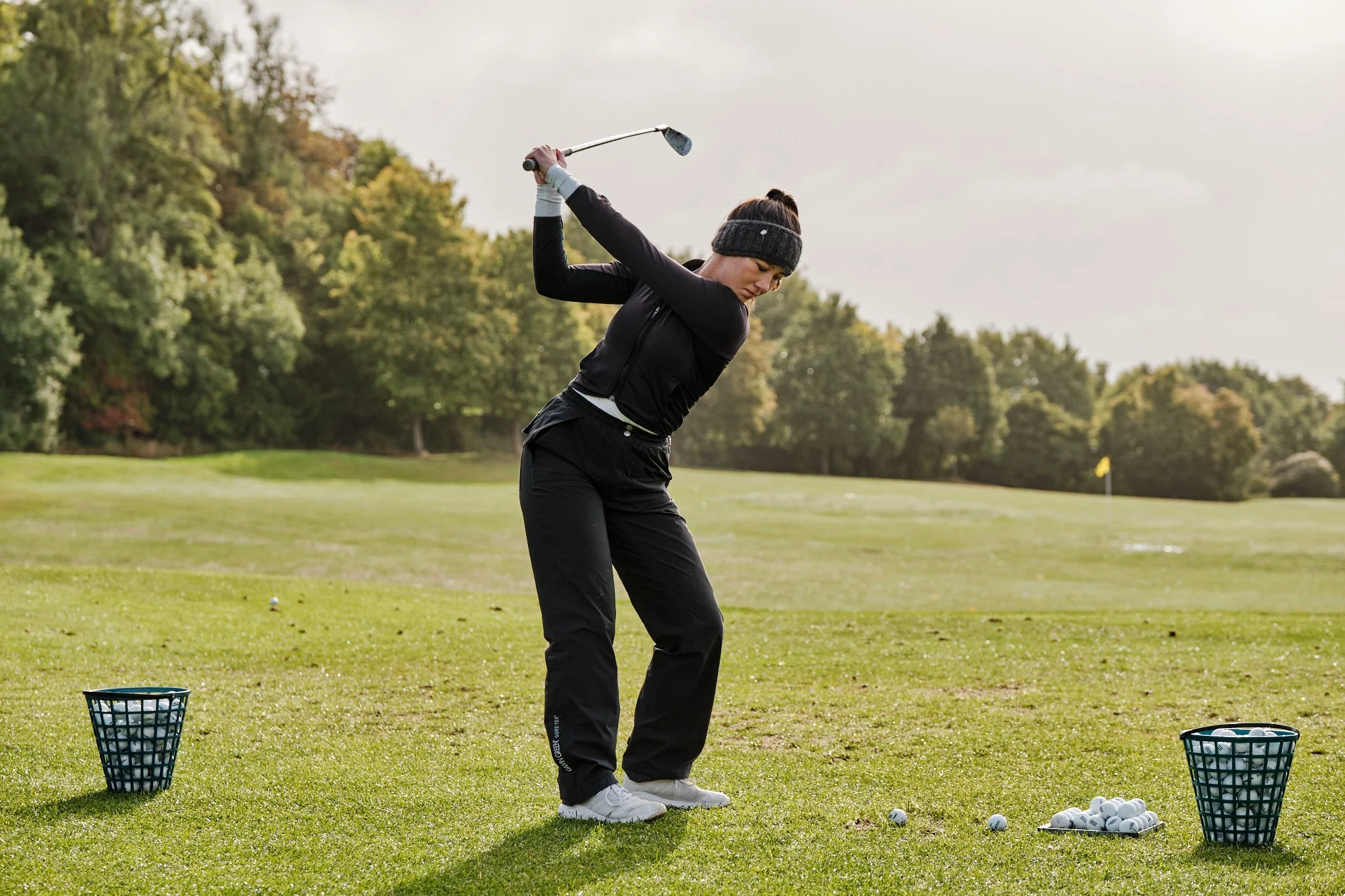 Woman in black athletic attire practicing golf swings on a golf course with baskets of golf balls nearby.