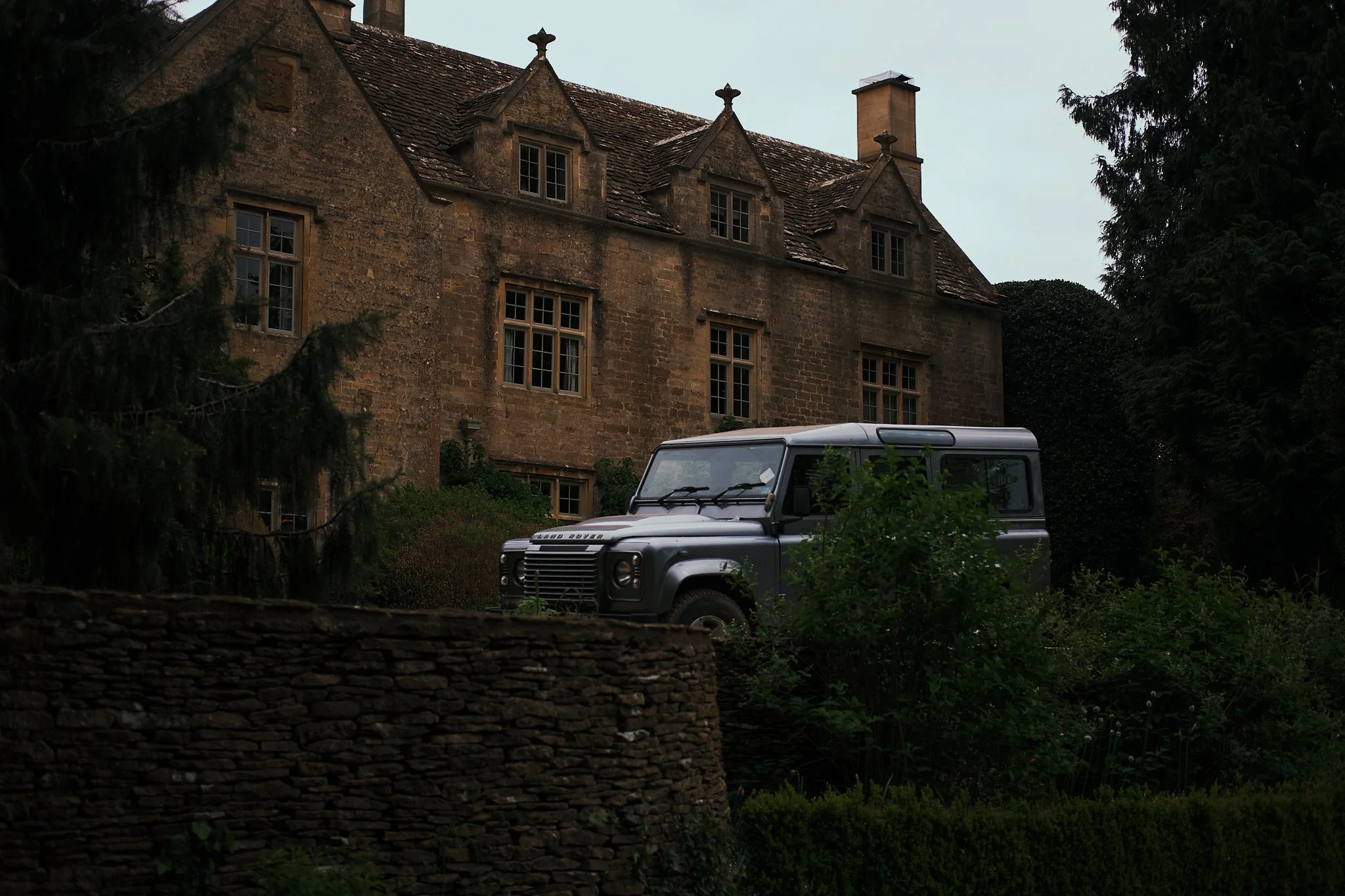 An old stone mansion with multiple windows and a tiled roof, partially obscured by trees, with a gray Land Rover parked in front amidst dense greenery. Hotel interior photography at THE PIG-in the Cotswolds