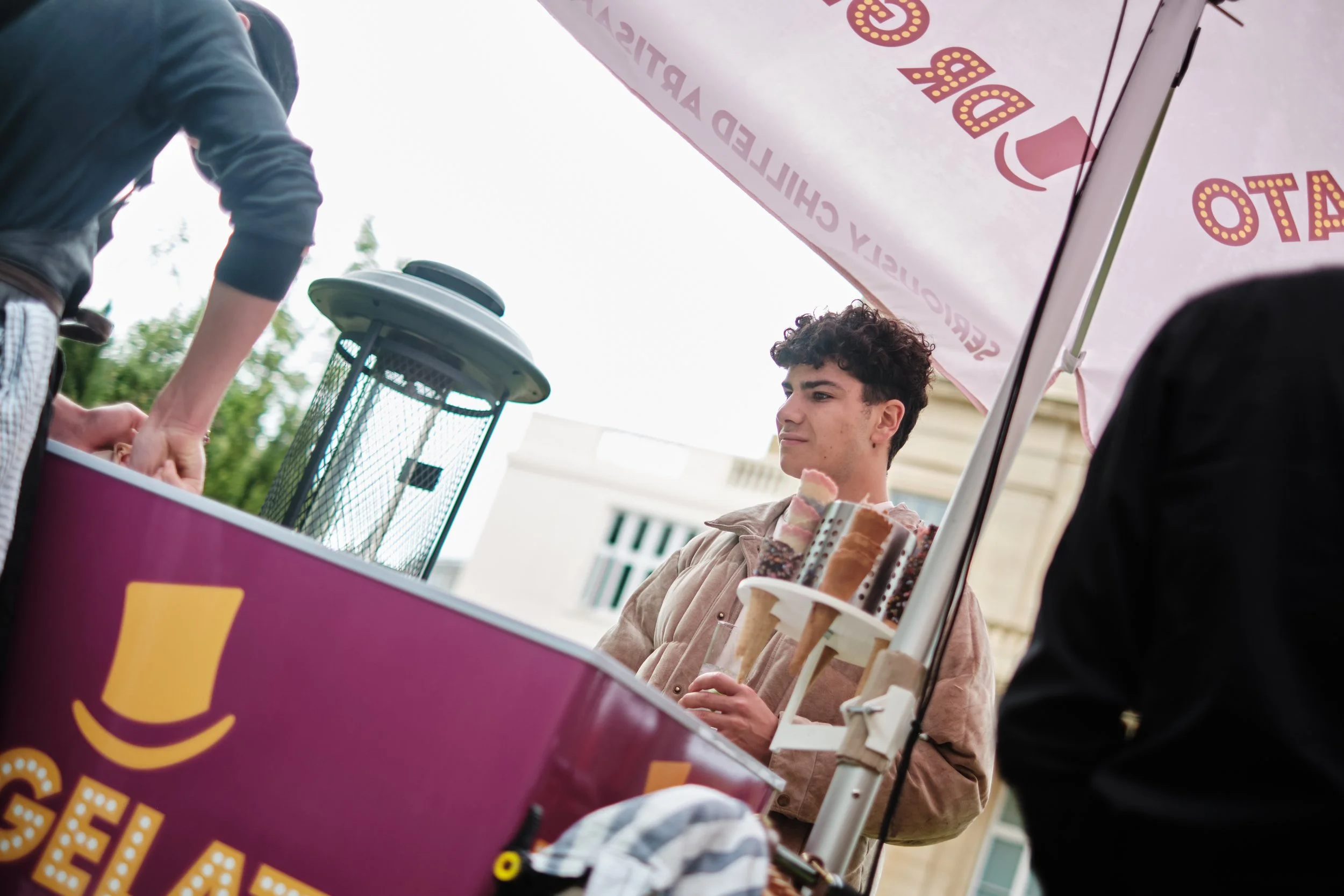 A young man at a gelato cart holding an ice cream cone, seen from a low angle with a partial view of an umbrella and another person handing him the ice cream.
