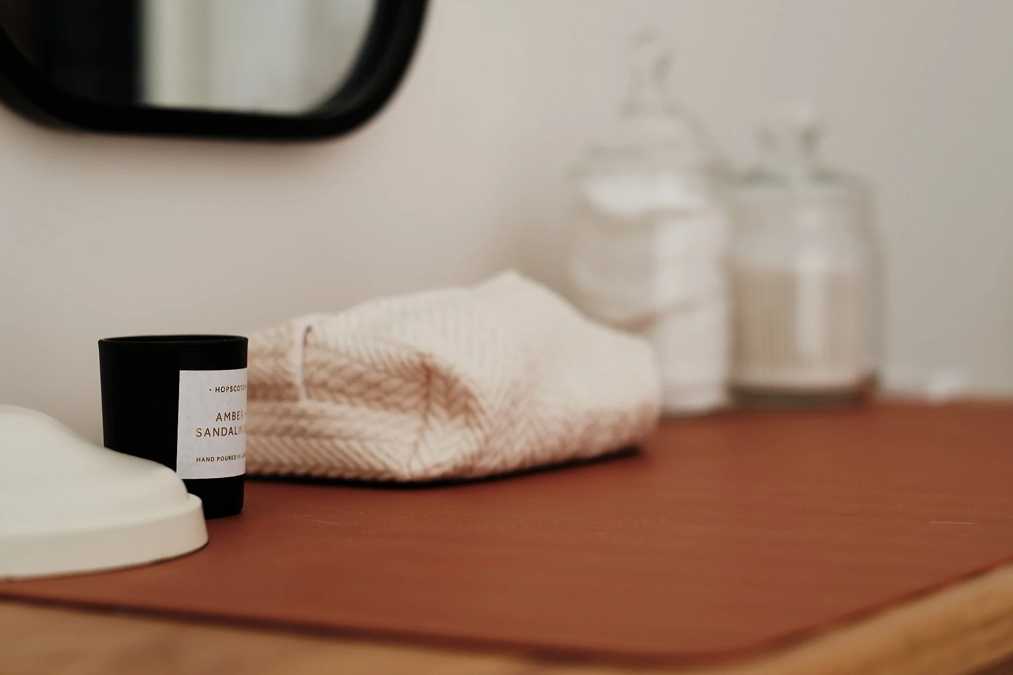 Close-up of a wooden surface with a black candle labeled 'Amber Sandalwood,' a folded pink cloth, and glass jars in the background, partially visible.