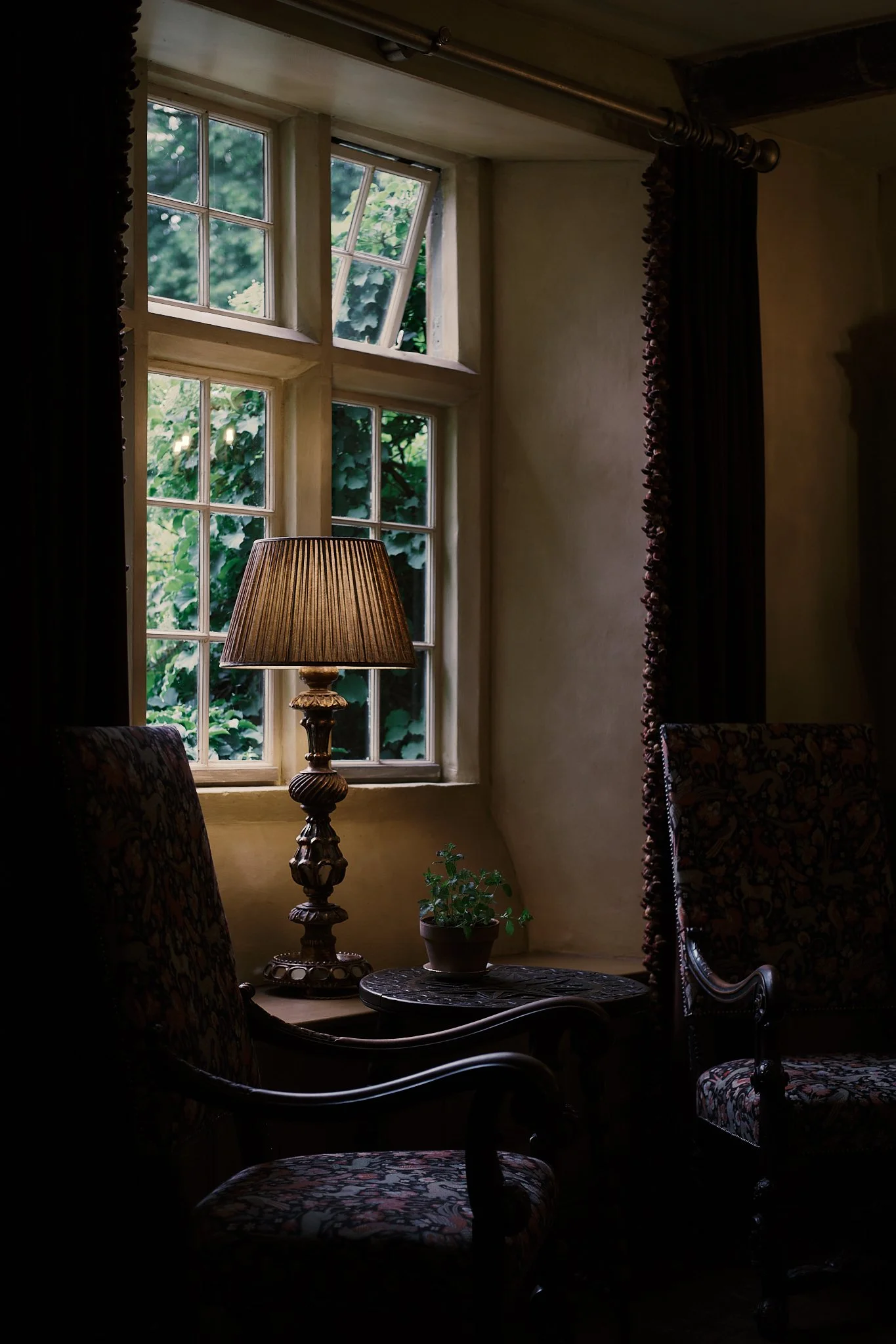 A cozy corner of a room with a window overlooking greenery, a table with a potted plant, and a lit table lamp between two patterned armchairs. Hotel interior photography at THE PIG-in the Cotswolds
