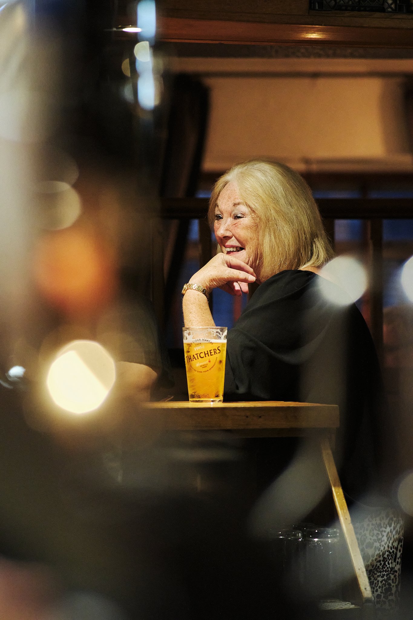 A smiling elderly woman seated at a bar with a glass of beer labeled 'Thatchers' in front of her, in a dimly lit pub or restaurant.
