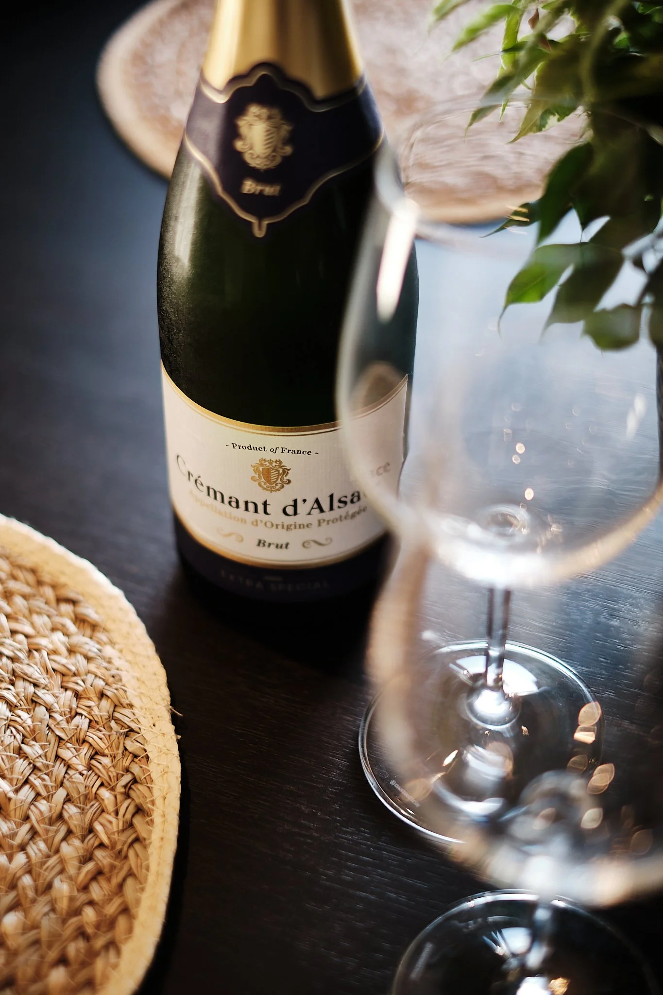 A bottle of Crémant d'Alsace sparkling wine, two empty wine glasses, a woven placemat, and a plant on a dark wooden table.