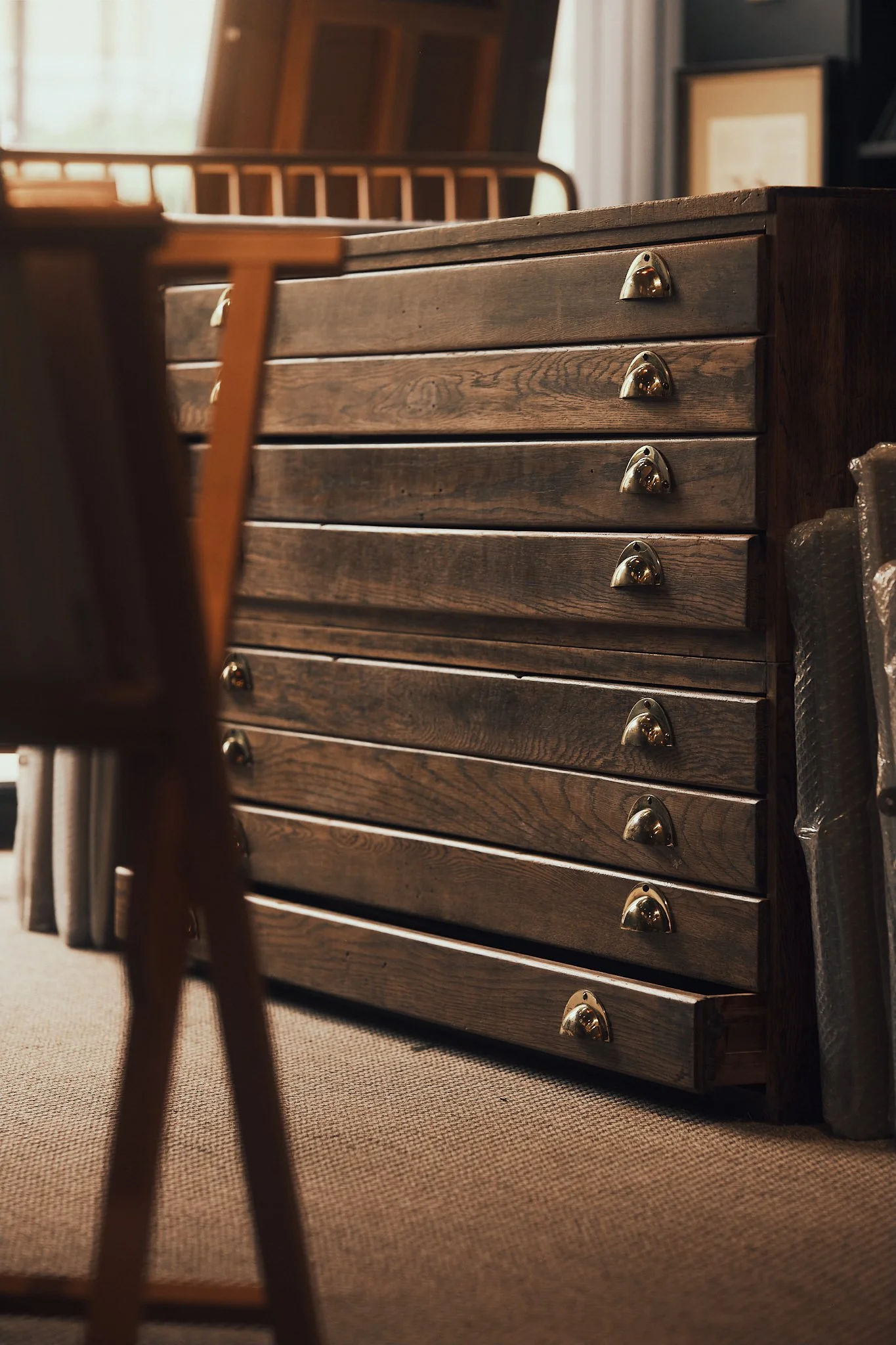 A vintage wooden filing cabinet with multiple drawers, brass handles, situated in a well-lit room with other furniture and framed pictures.