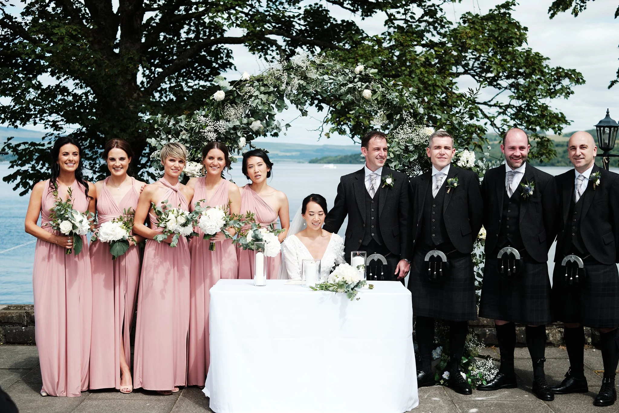Group of nine people at a wedding ceremony outdoors by a lake, with a large tree and decorated floral arch in the background. Six women on the left and three men on the right, all dressed formally, smiling.