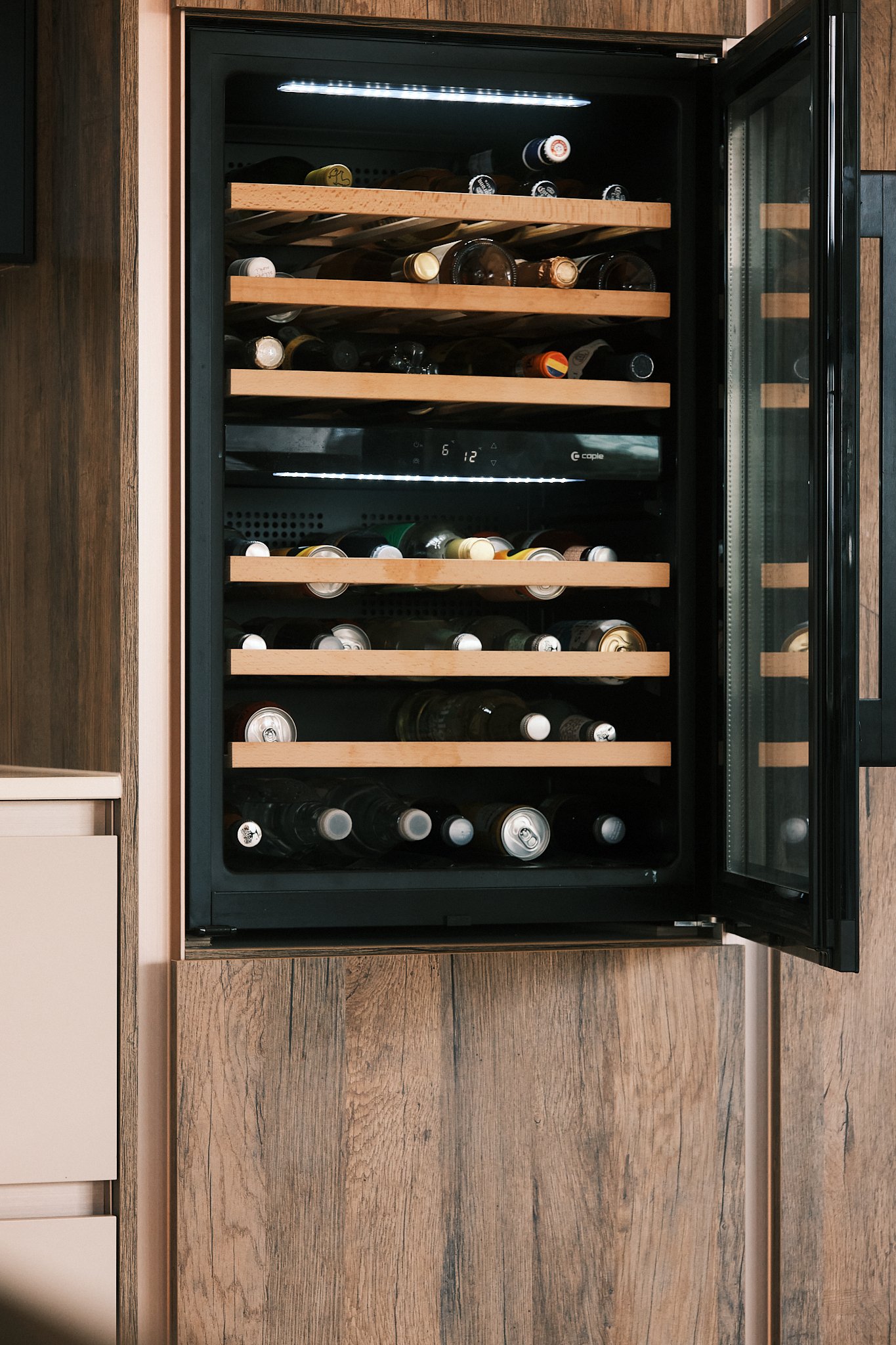 A wine cooler stocked with various bottles of wine on wooden shelves inside, built into a wooden-paneled wall.