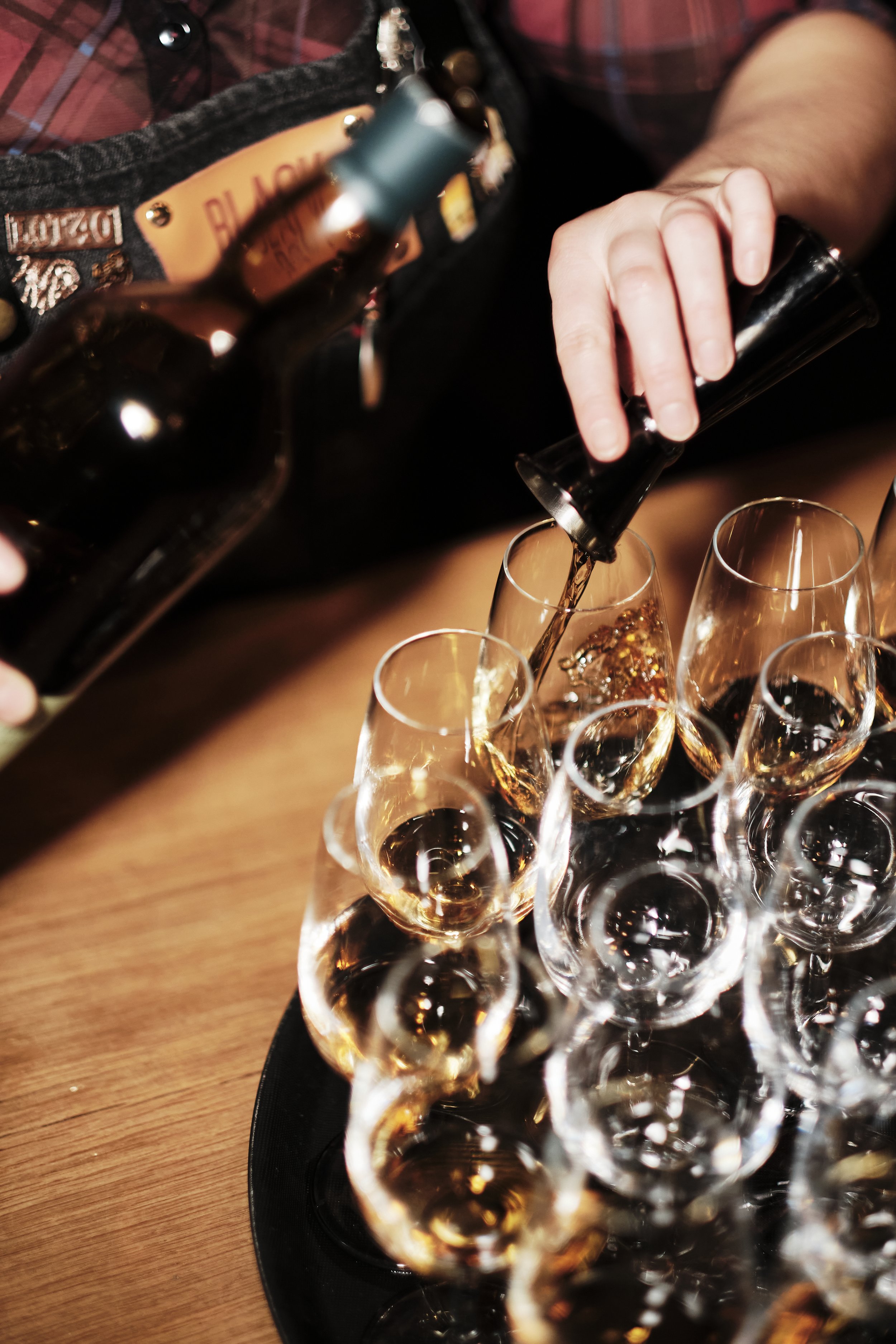 Person pouring a drink into a glass of whiskey or wine on a table surrounded by other glasses.