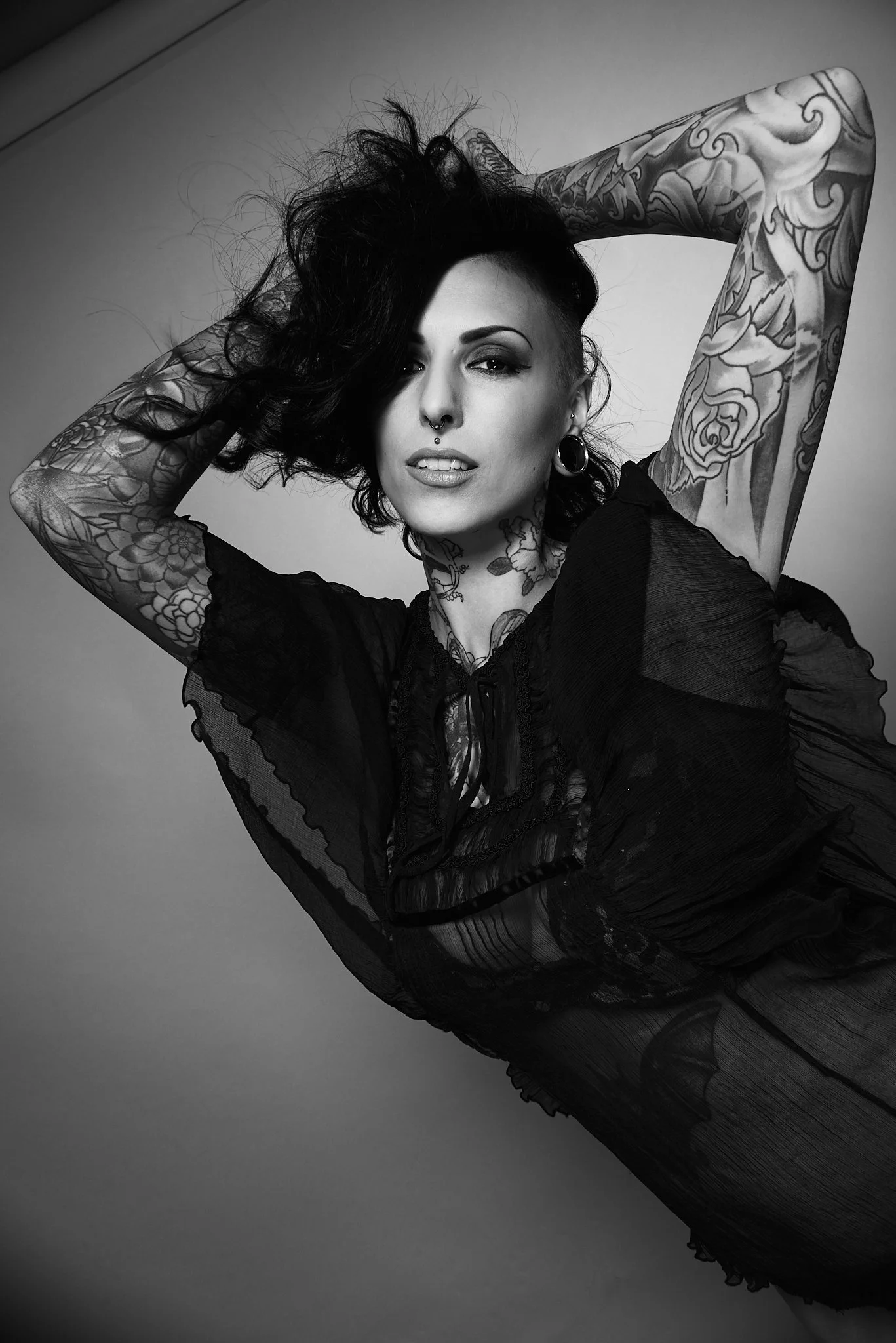Black and white portrait of a woman with tattoos, piercings, and dark hair posing with arms above her head, wearing a sheer black top.