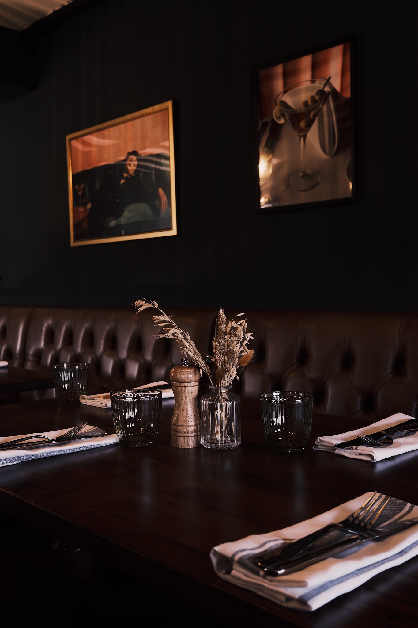 A restaurant table with napkins, forks, and glasses, a vase with dried flowers, and wall art including a portrait and a drink