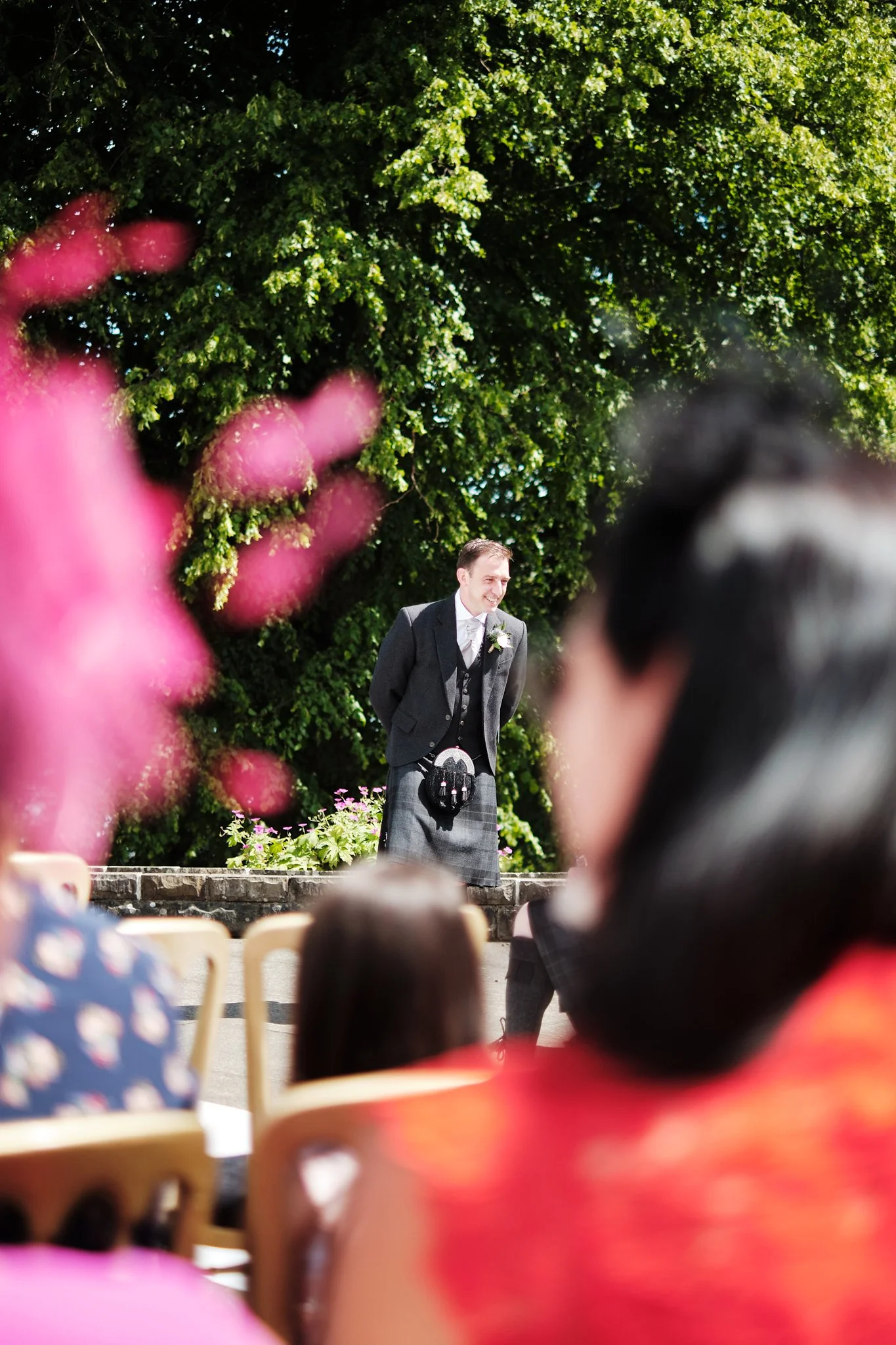 A man in a gray suit and kilt standing outdoors during a wedding ceremony, smiling, with a large green tree in the background, as children and others sit in front, with blurred pink flowers in the foreground.