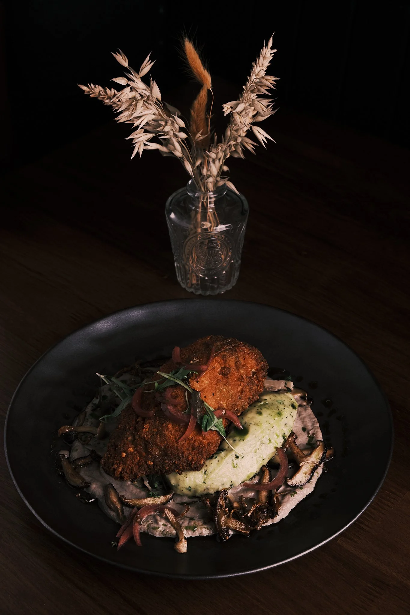 A black plate with breaded fried fish fillets topped with herbs, served over mashed potatoes with mushrooms, onions, and a light sauce, on a dark wooden table with a vase of dried flowers in the background.