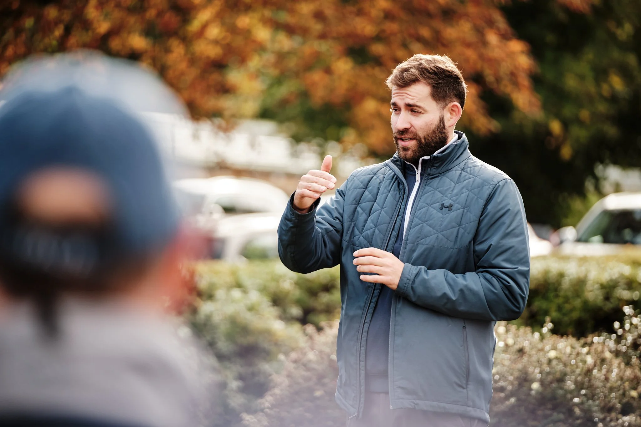 A man with a beard and short hair wearing a blue quilted jacket explaining or talking to someone outdoors during fall, with trees with red and orange leaves in the background.