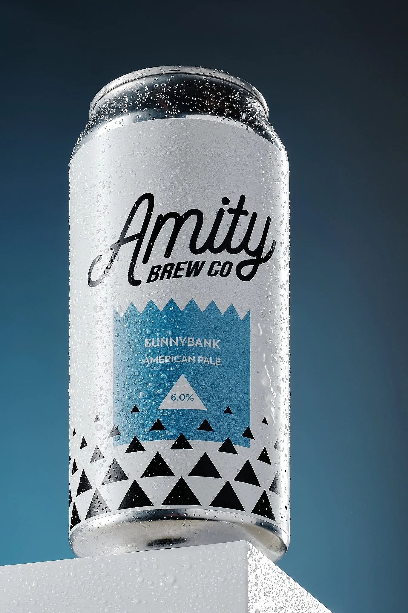 A can of Amity Brew Co's Sunnybank American Pale Ale with condensation droplets on its surface, placed on a white surface against a dark blue background.