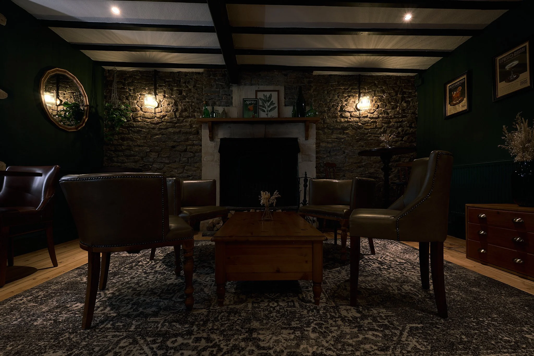 A cozy living room with dark green walls, a stone fireplace, leather armchairs, a wooden coffee table, and warm wall-mounted lights.