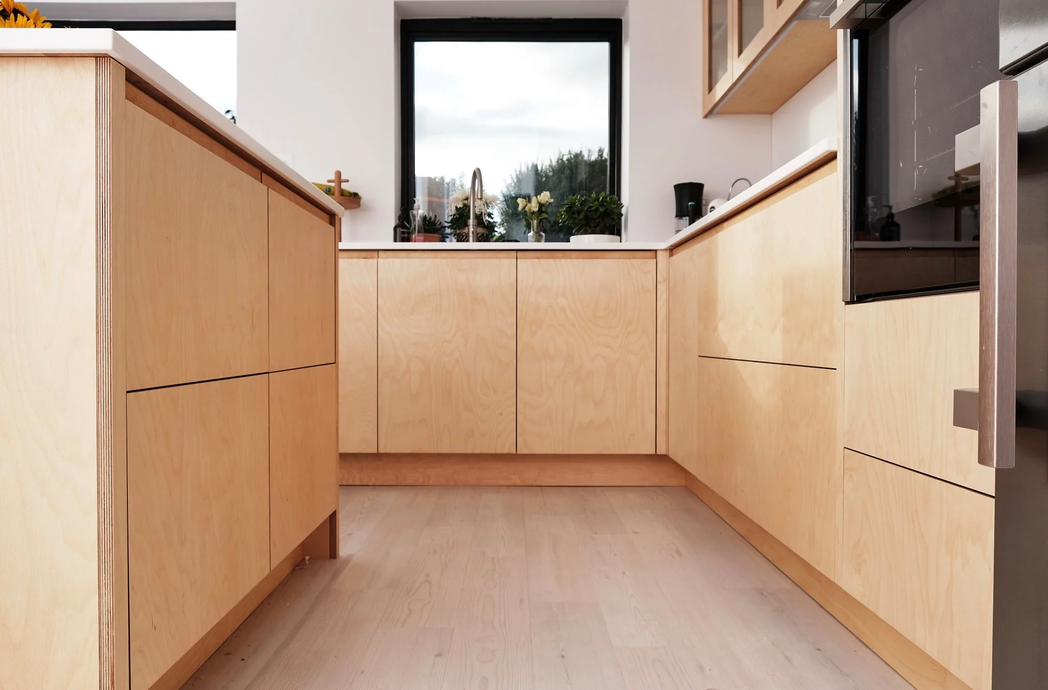 A modern kitchen with light wooden cabinets, a white countertop, and a window above the sink with plants and flowers, and a view of the outdoors.
