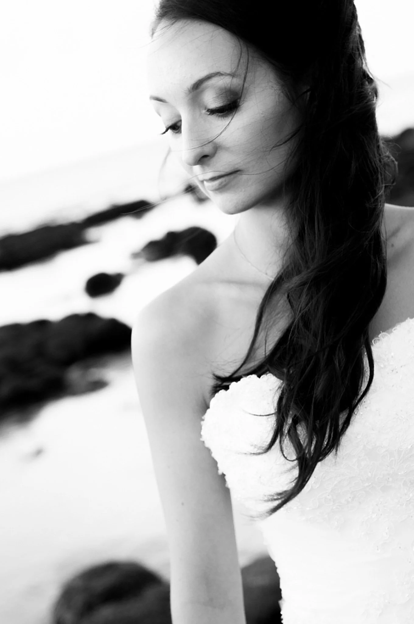 A black and white photo of a woman with long, wavy hair looking down, standing outdoors on a beach with rocks in the background.
