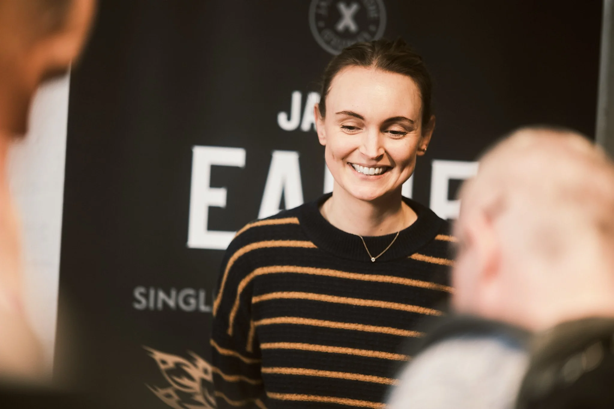 A smiling woman with dark hair in a striped black and orange sweater, engaging with two people in the foreground, with a black background displaying the word 'FAITH'.