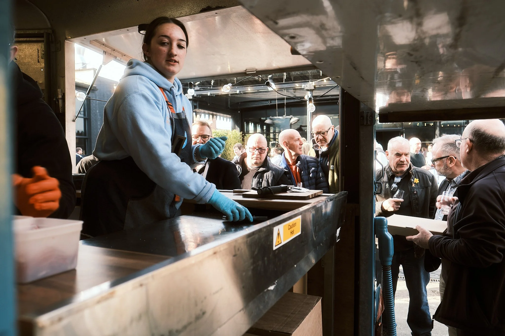 A young woman with brown hair wearing a blue hoodie, black apron, and blue gloves working behind a food stall counter, with a group of men in jackets and glasses waiting and talking outside in the background.
