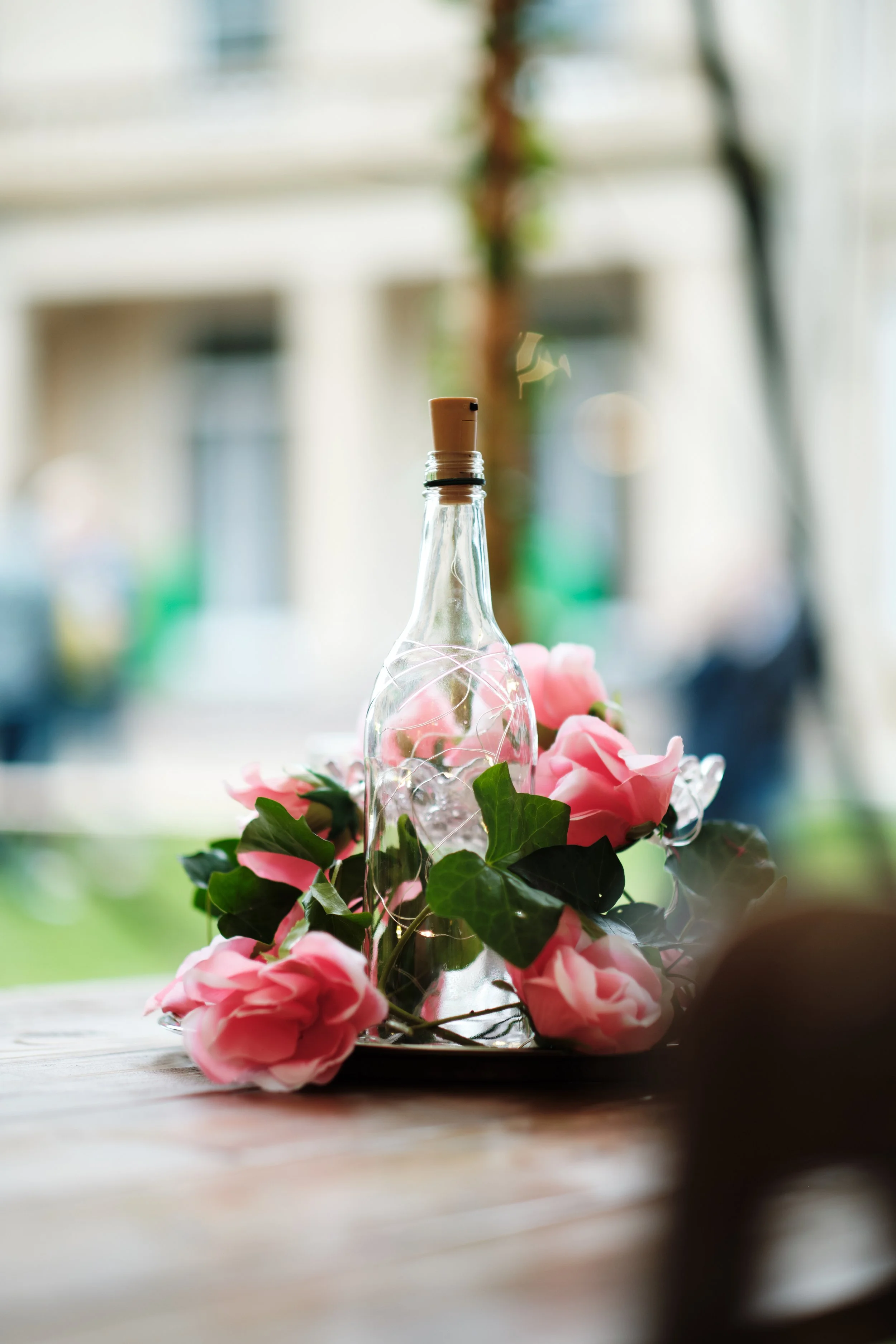 A clear glass bottle with a cork stopper, surrounded by pink flowers and green ivy leaves, placed on a wooden table near a window.